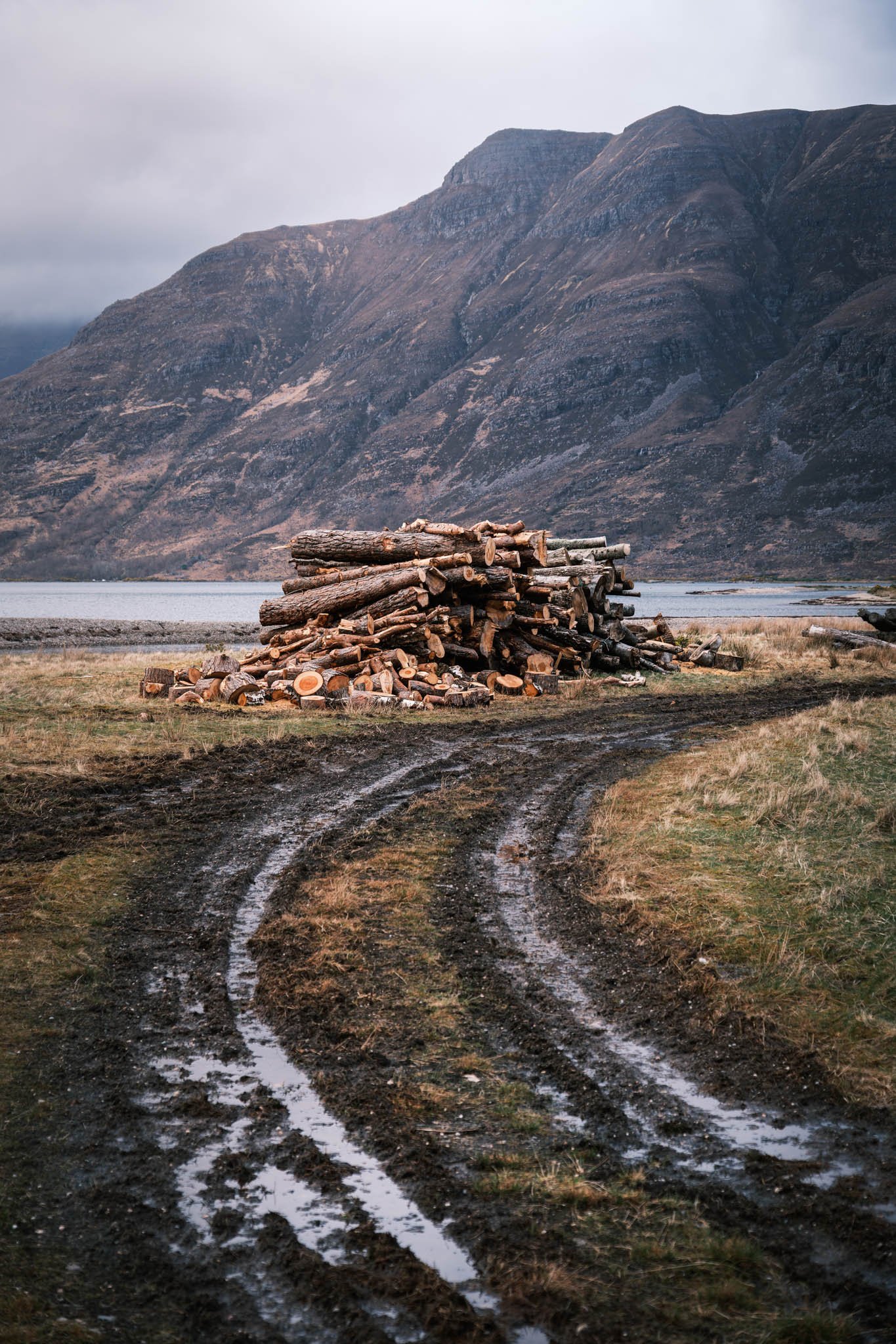 Highland - Scotland - Montagnes brunes avec peu de végétation, étendue d'eau au premier plan, piste boueuse avec de la boue ou de l'eau, ciel nuageux.