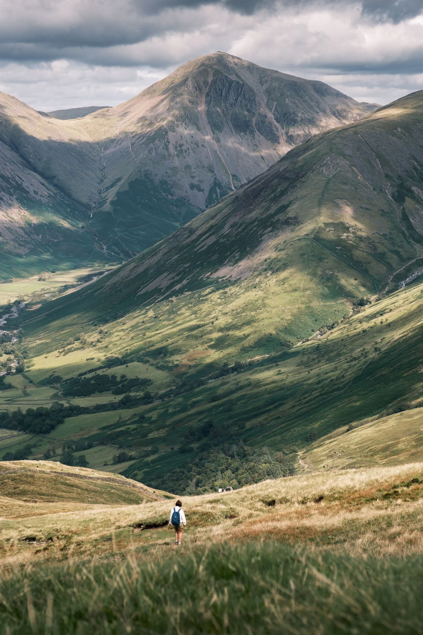 Lake District - Personne marchant dans une vallée verdoyante entourée de montagnes escarpées sous un ciel nuageux.  Reportage outdoor, mise en valeur du territoire et tourisme durable par Guillaume Donsimoni.