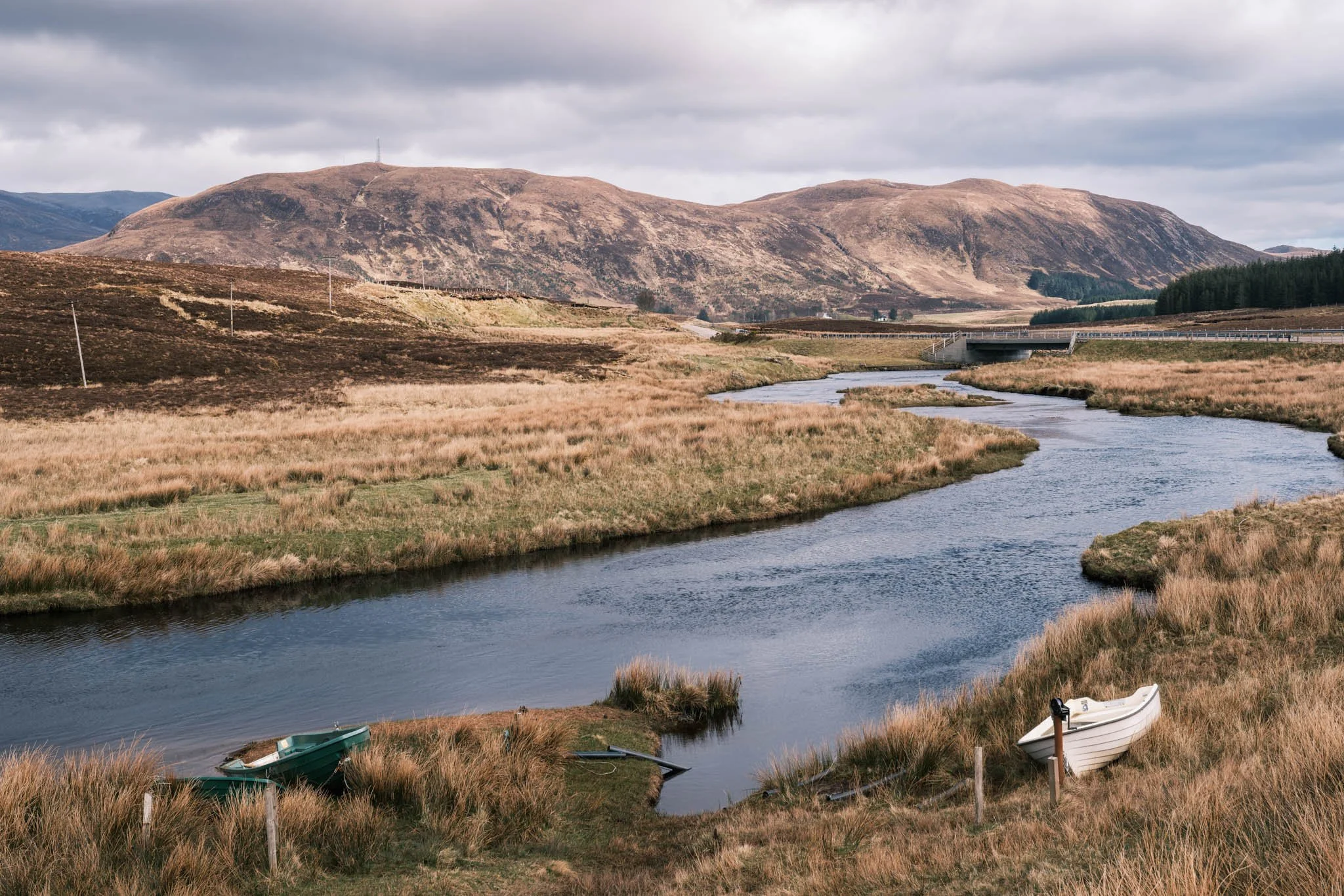 Highland - Scotland - Paysage avec ruisseau, montagnes brunes, nuages gris, avec deux petits bateaux à la berge.