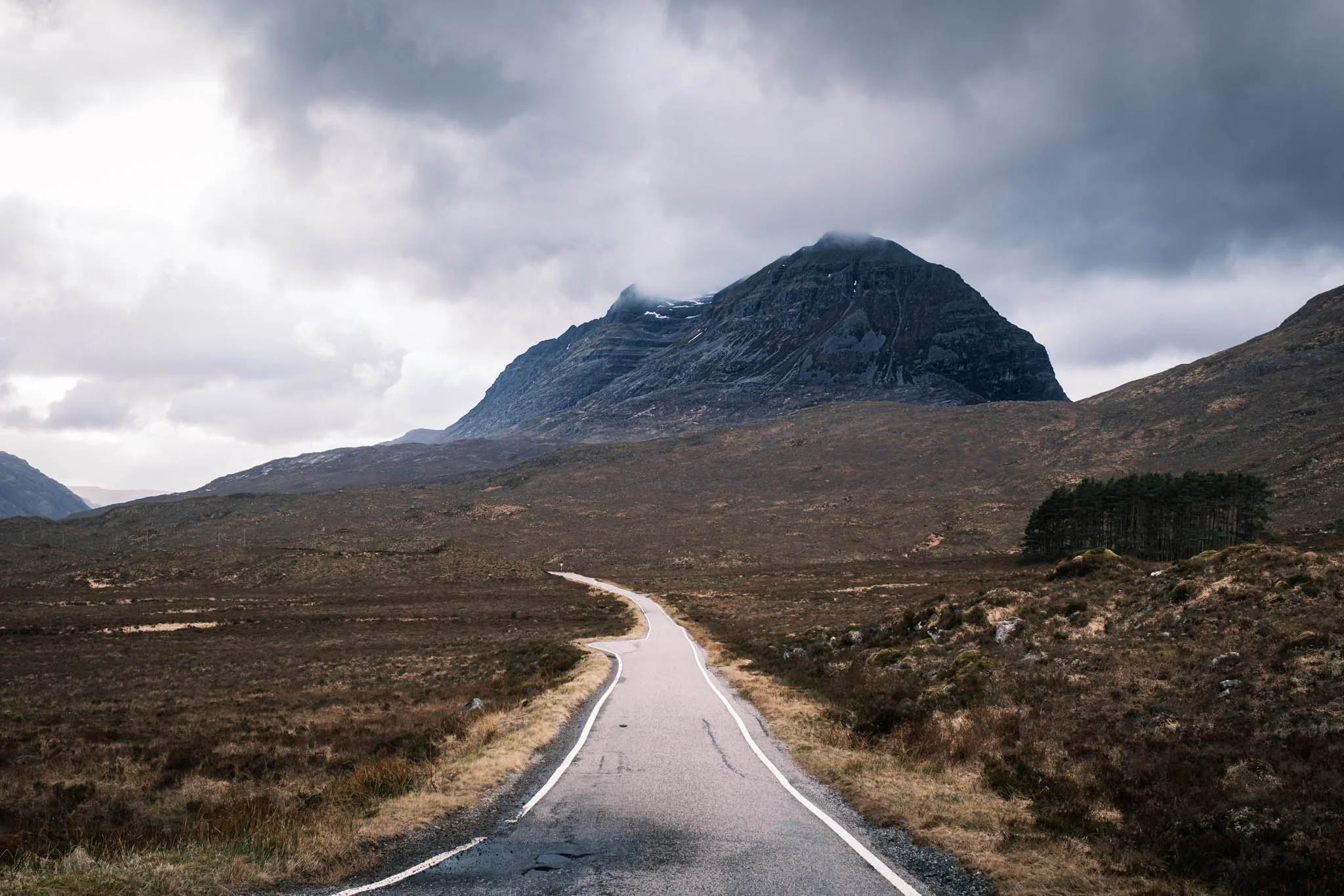 Highland - Scotland - Une route sinueuse traverse un paysage de landes avec une montagne escarpée en arrière-plan, sous un ciel nuageux.