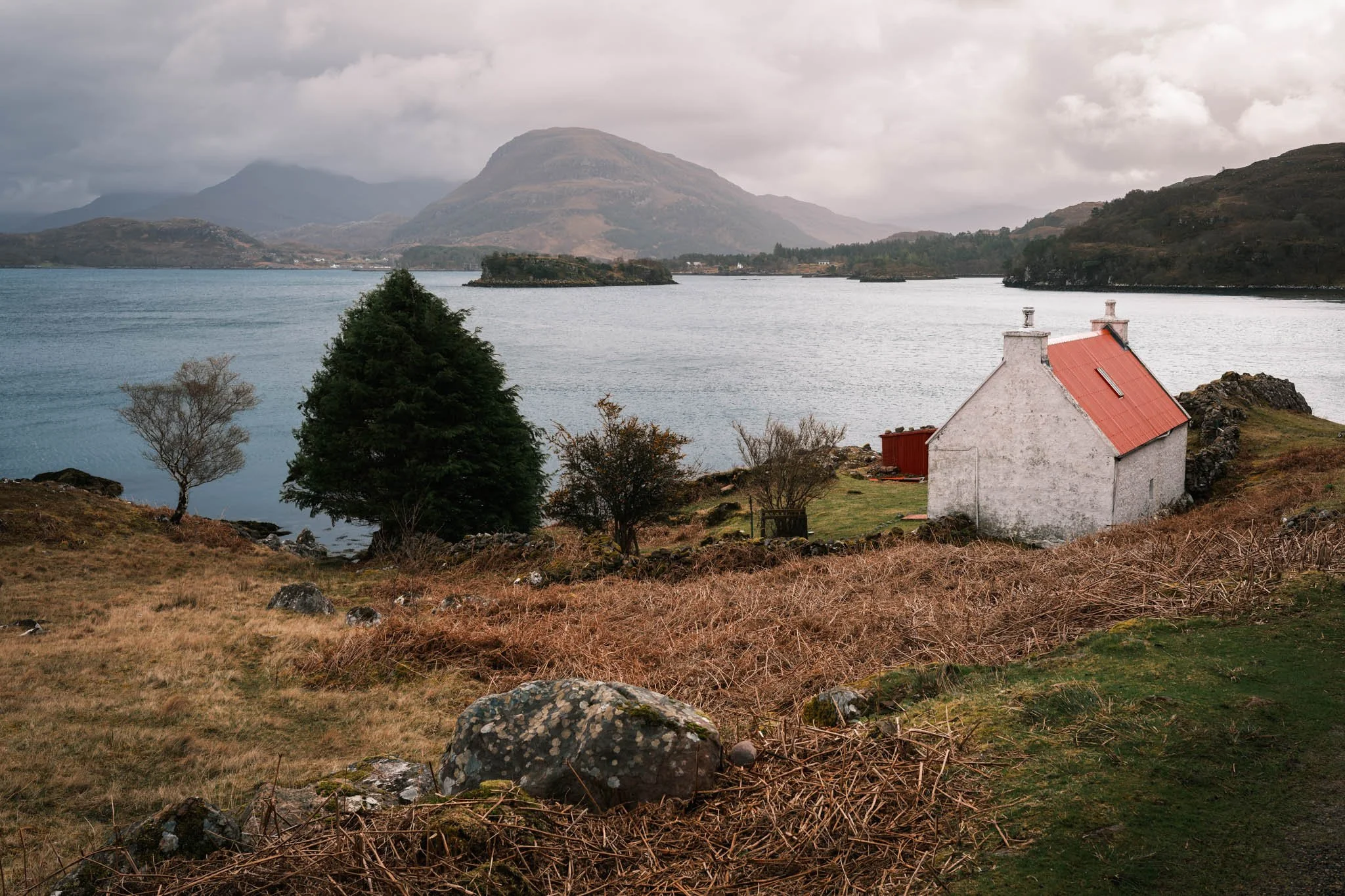Highland - Scotland - Un Loch entouré de montagnes avec un ciel nuageux; en premier plan, une maison blanche avec un toit en tôle rouge, des arbres et des rochers.