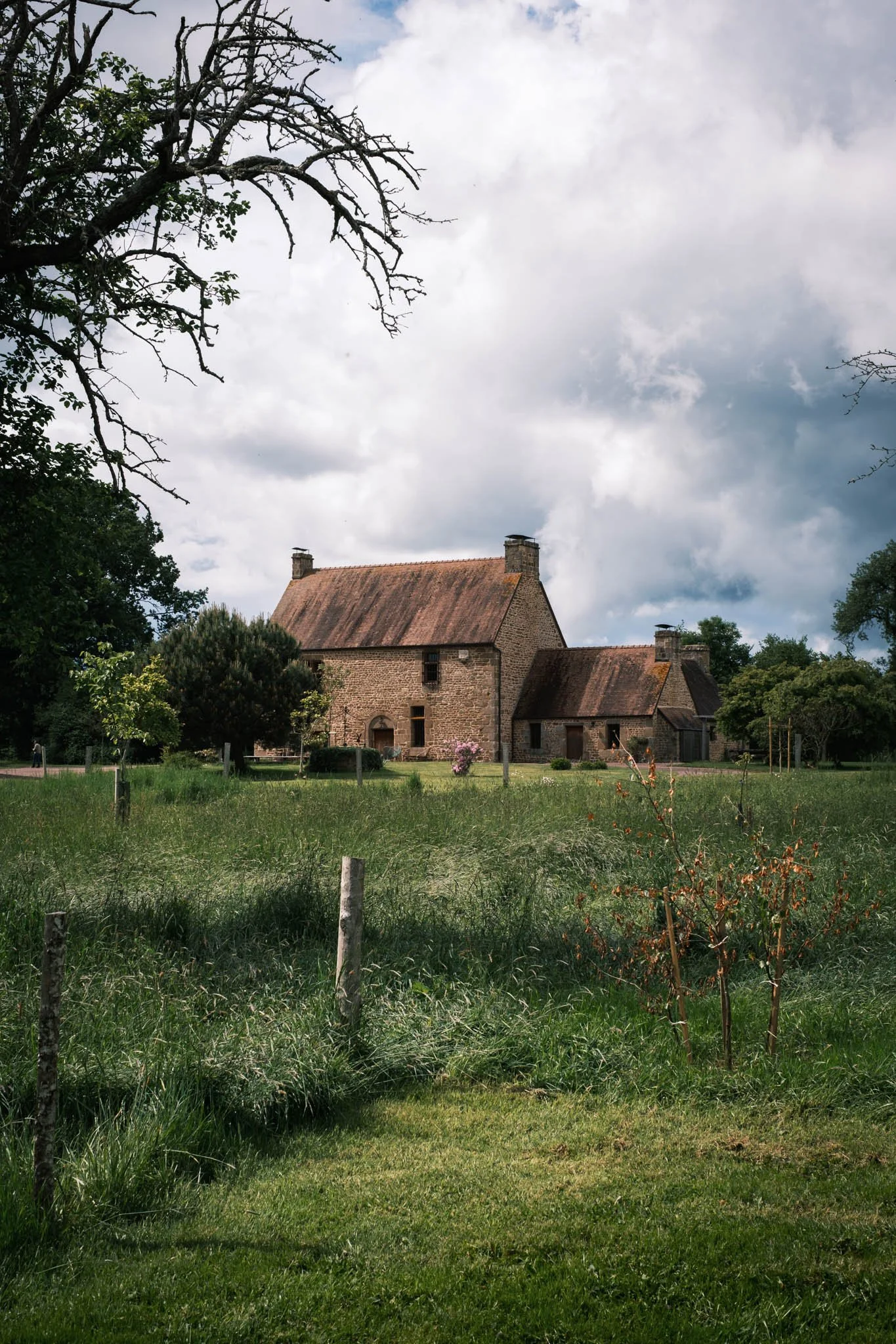 Suisse Normande - Une maison ancienne en pierre avec un toit en tuiles rouges, entourée de arbres et de pelouses vertes, sous un ciel partiellement nuageux.