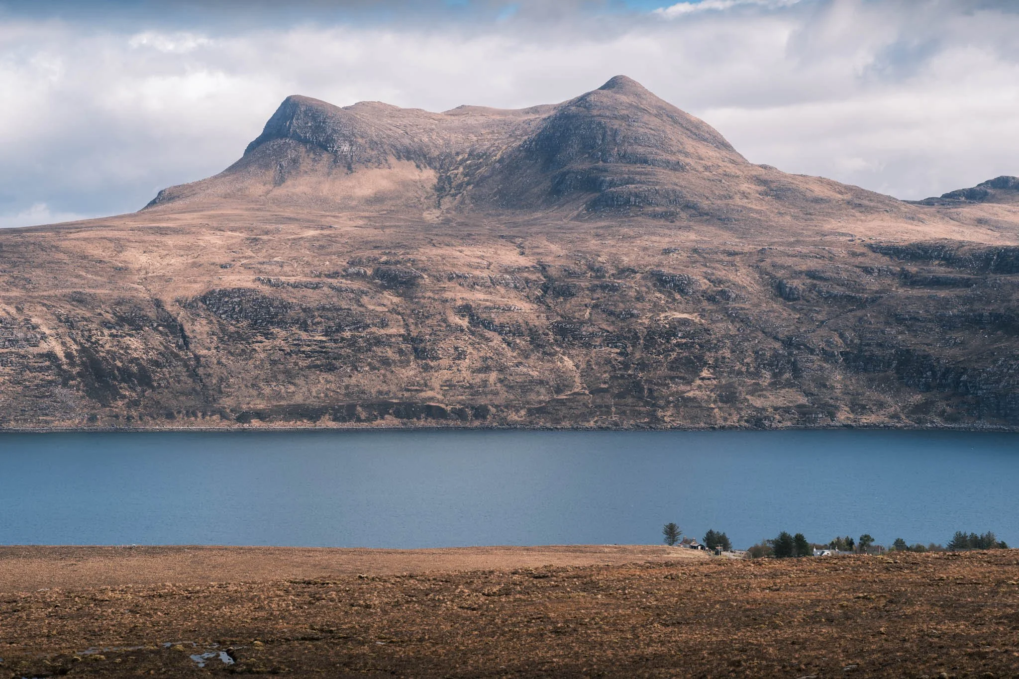 Highland - Scotland - Montagne rocheuse avec un sommet arrondi surplombant un Loch bleu, avec un petit bâtiment et des arbres au premier plan.