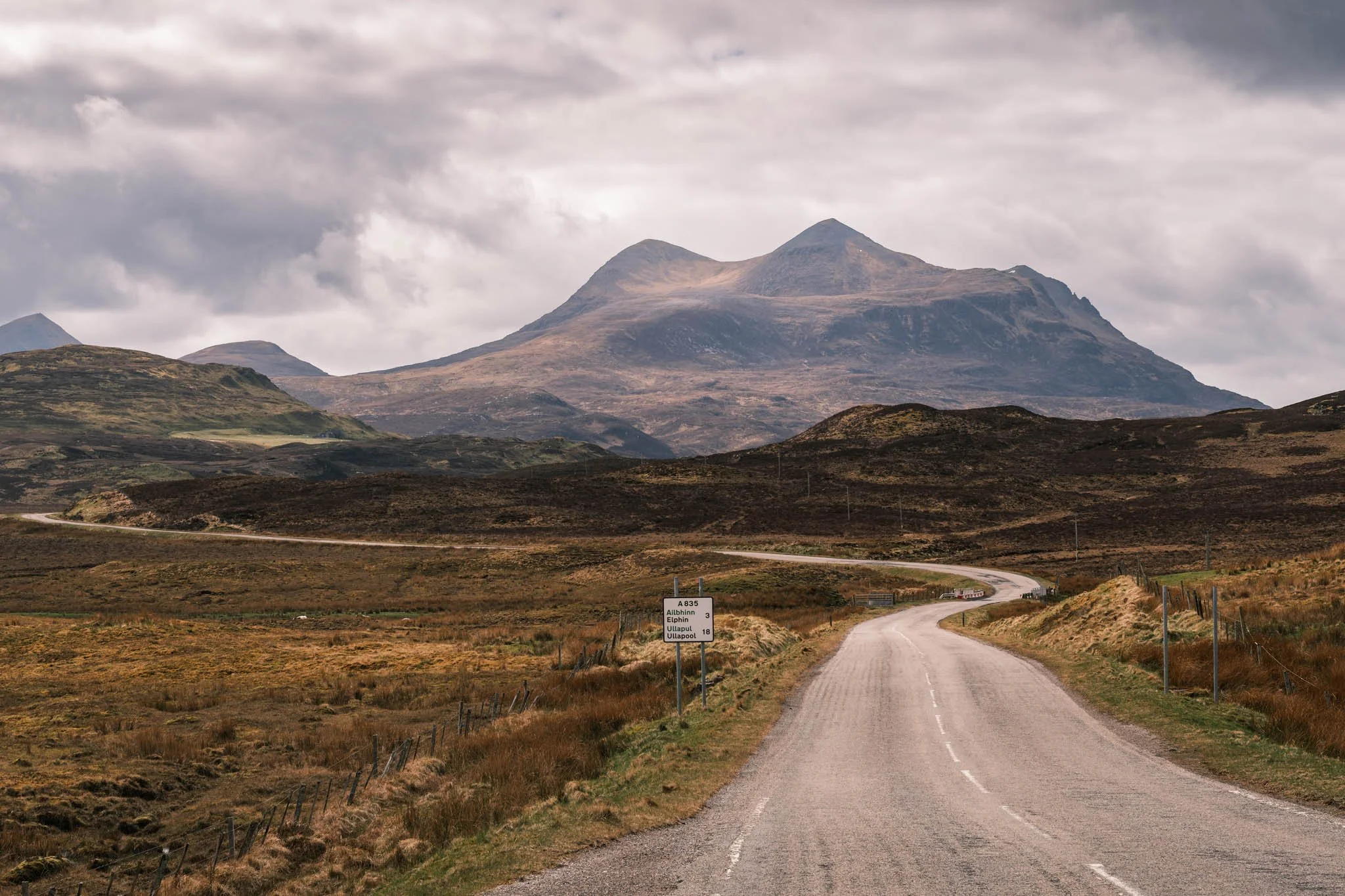 Highland - Scotland - Une route rurale serpente à travers un paysage de collines et de montagnes sous un ciel nuageux.