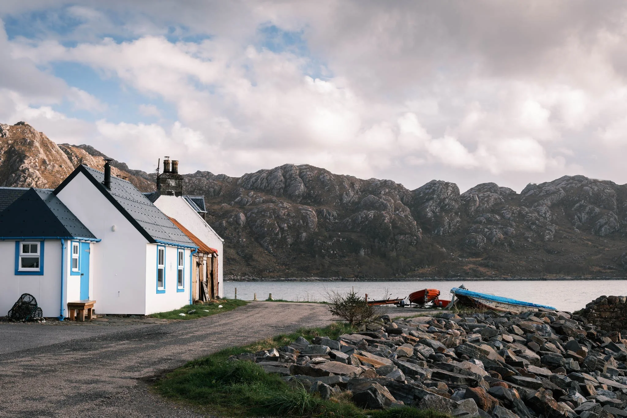 Highland - Scotland - Maisons blanches avec toits en tôle gris, un bateau et des rochers au bord de l'eau, montagnes en arrière-plan, ciel nuageux.