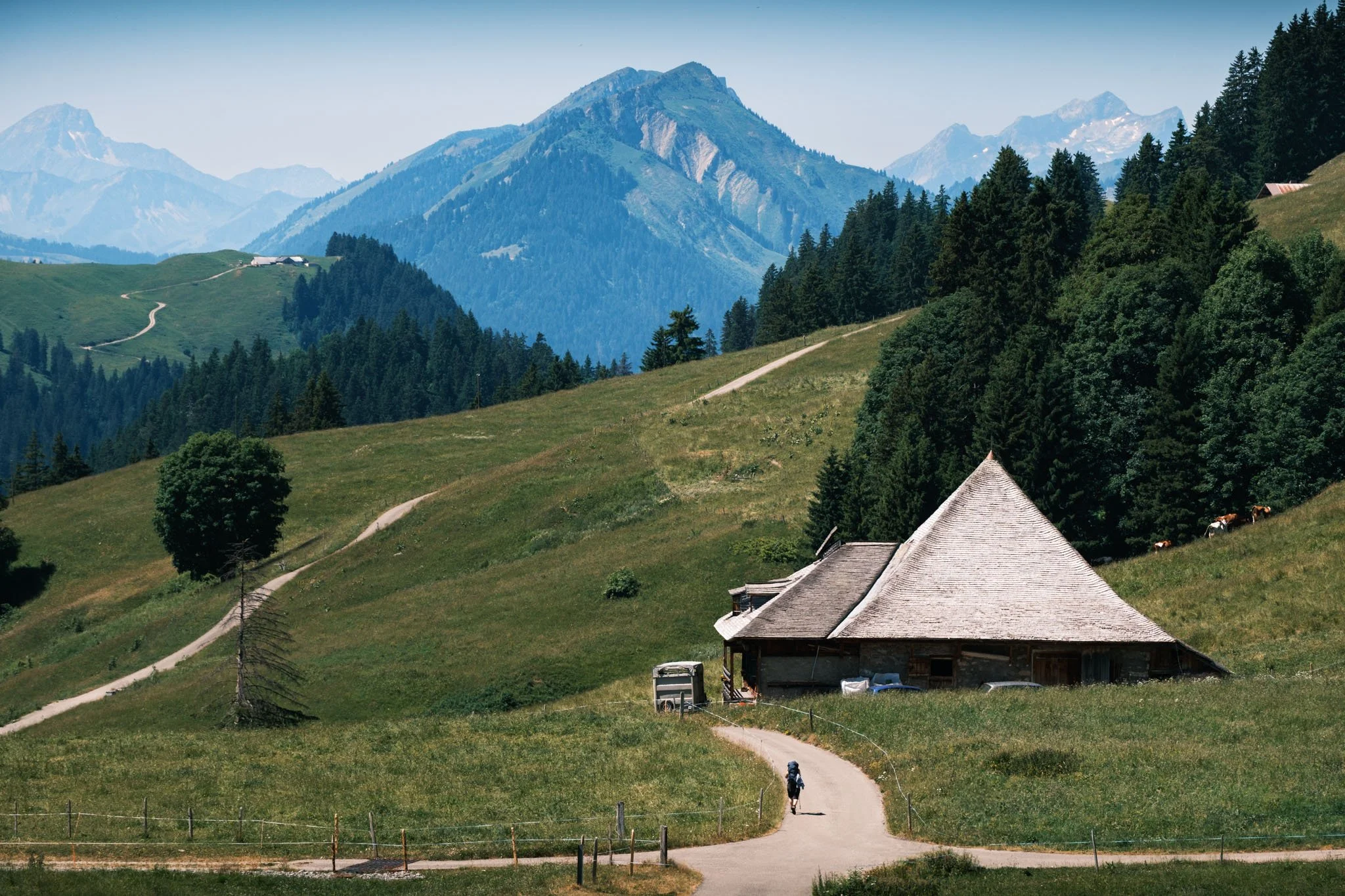 Paysage de montagne avec une maison en bois à toit pyramidal, sentier de terre, personne à vélo, arbres et montagnes en arrière-plan. Reportage outdoor, mise en valeur du territoire et tourisme durable par Guillaume Donsimoni