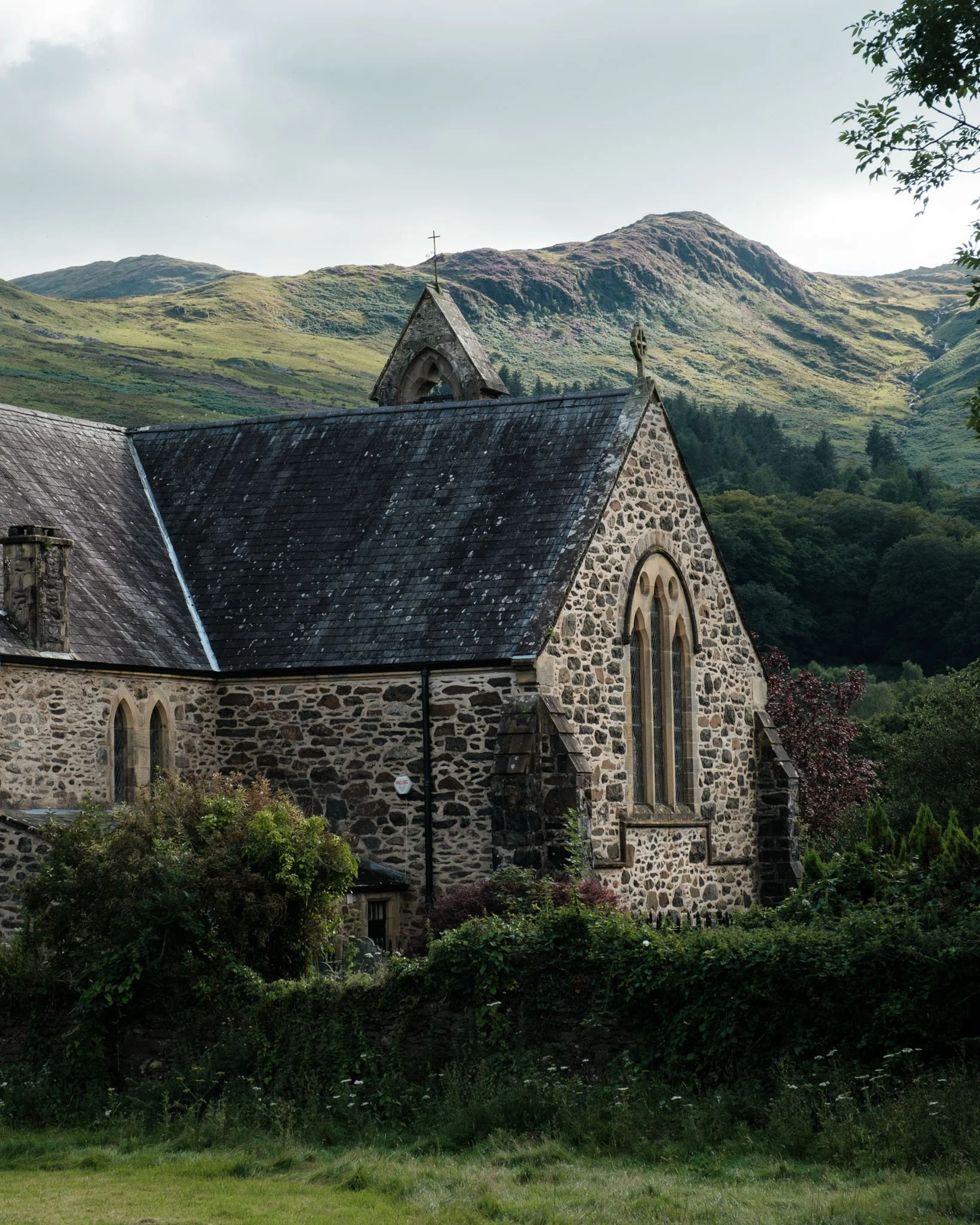 Wales - An old stone church with a bell tower, situated amidst dense vegetation, with green mountains in the background.