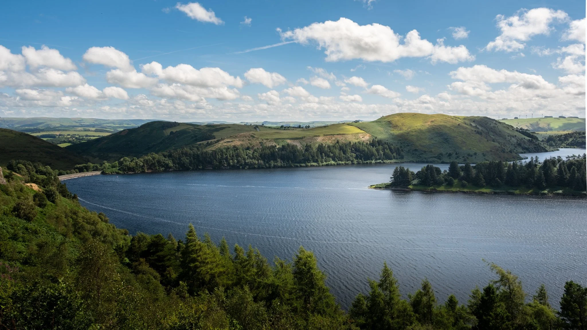 Lake surrounded by green hills under a partially cloudy sky. Outdoor reportage, regional promotion, and sustainable tourism by Guillaume Donsimoni.