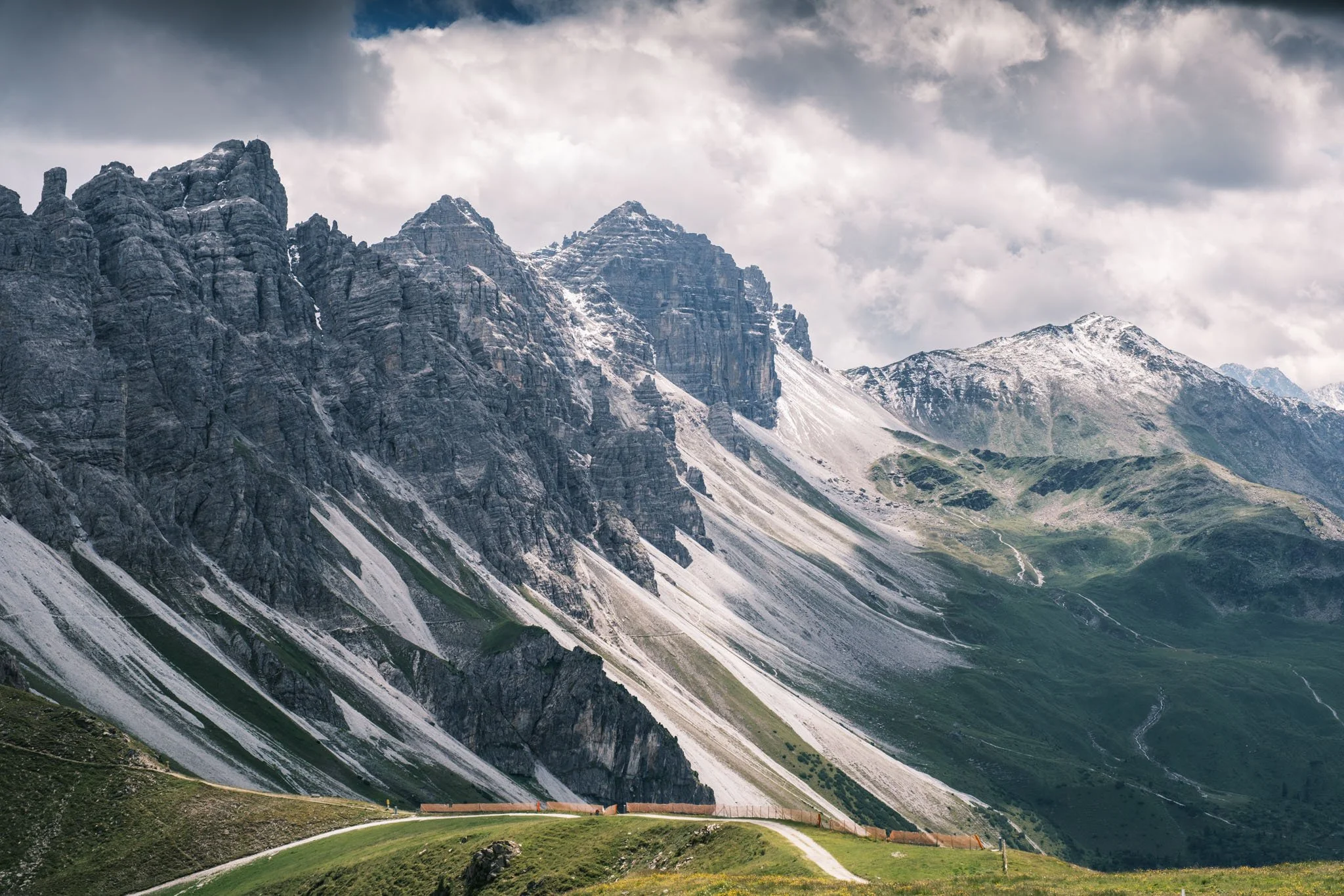 Tyrol - Montagnes imposantes avec sommets enneigés, prairie verte et routes sinueuses en premier plan, ciel nuageux en arrière-plan. Reportage outdoor, mise en valeur du territoire et tourisme durable par Guillaume Donsimoni