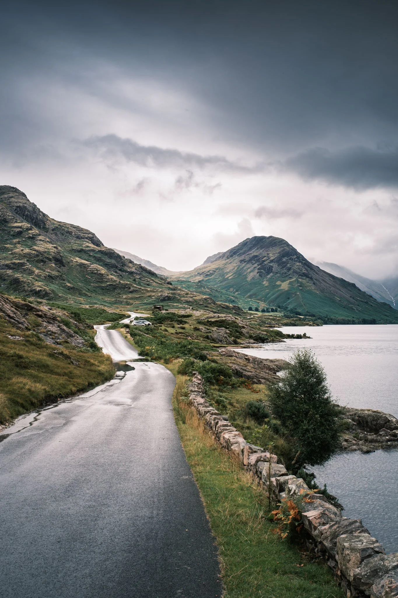 Lake District - Une route pavée longeant un lac de montagne, avec des montagnes vertes sous un ciel nuageux.  Reportage outdoor, mise en valeur du territoire et tourisme durable par Guillaume Donsimoni.