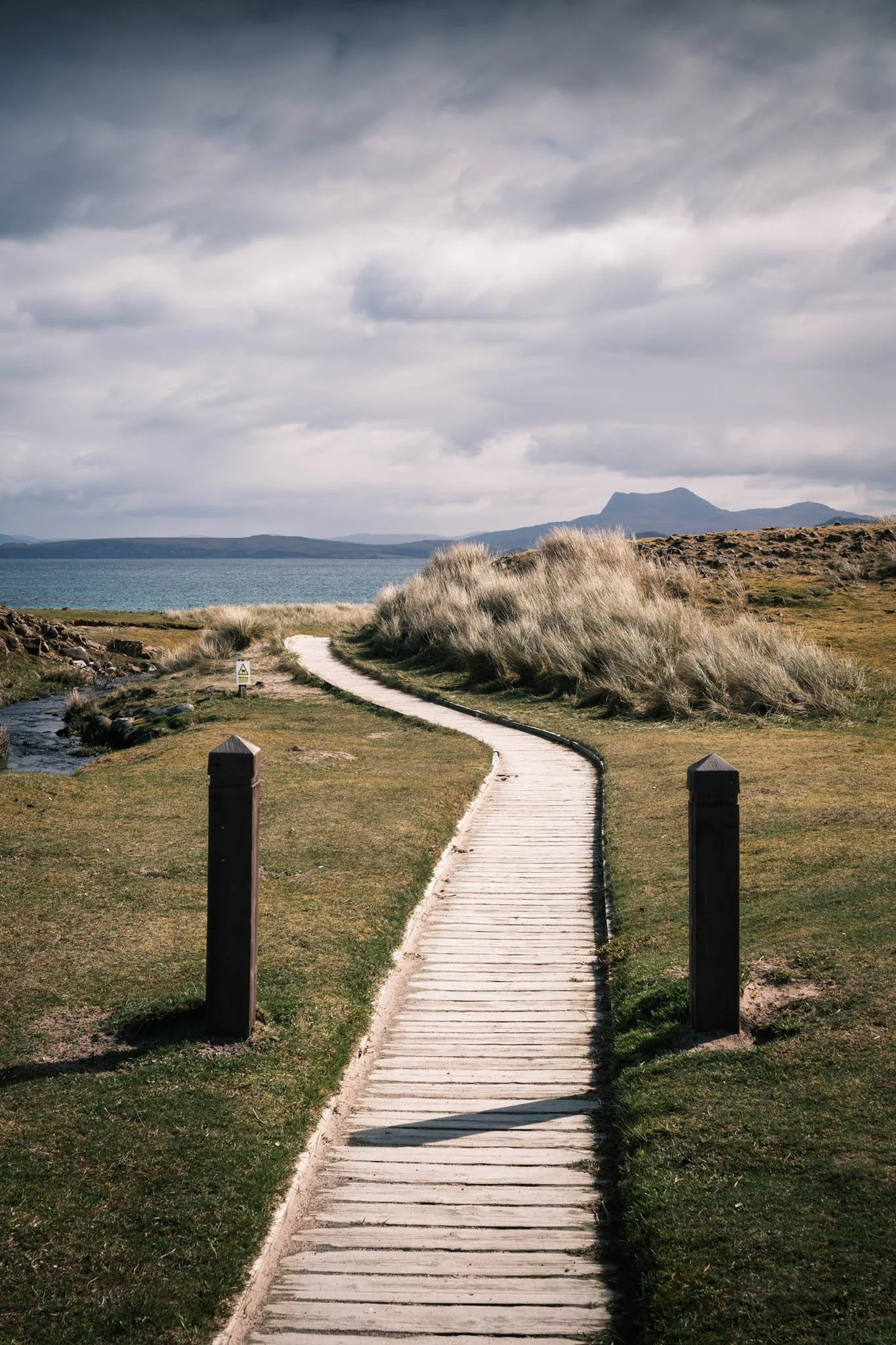 Highland - Scotland - Chemin en bois serpentant à travers un paysage naturel avec herbes hautes, un Loch et des montagnes au loin, sous un ciel nuageux.
