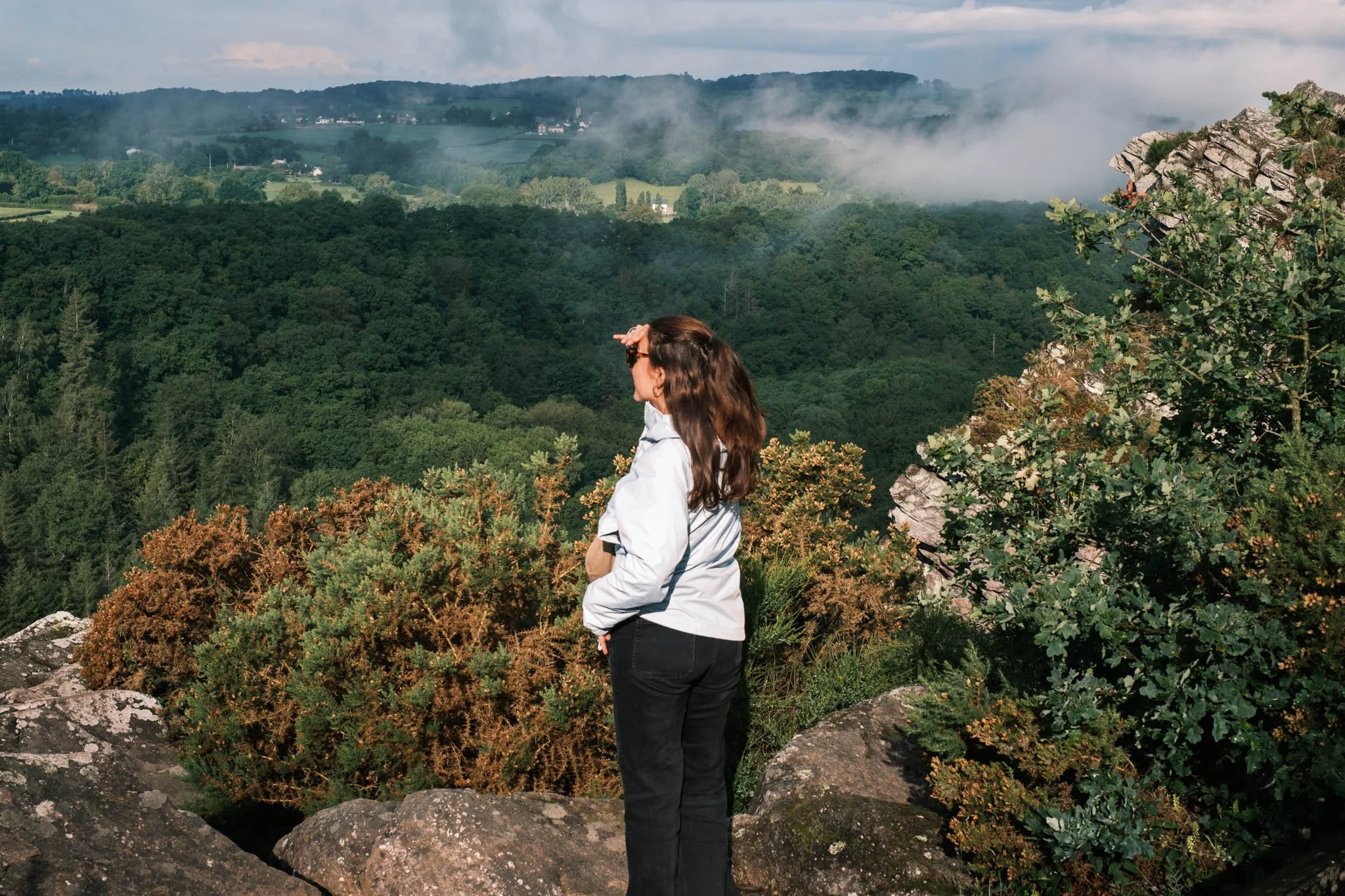 Suisse Normande - Une femme avec des lunettes regarde à l'horizon, debout sur un rocher, entourée de végétation luxuriante, avec des collines et de la brume en arrière-plan, sous un ciel nuageux.