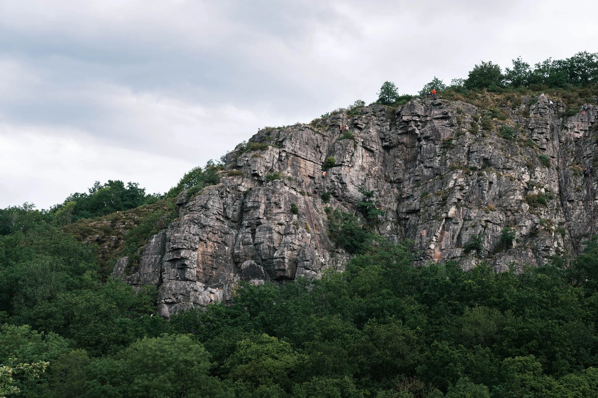 Suisse Normande - Massif rocheux avec des grimpeurs en escalade, entouré de verdure et sous un ciel nuageux.
