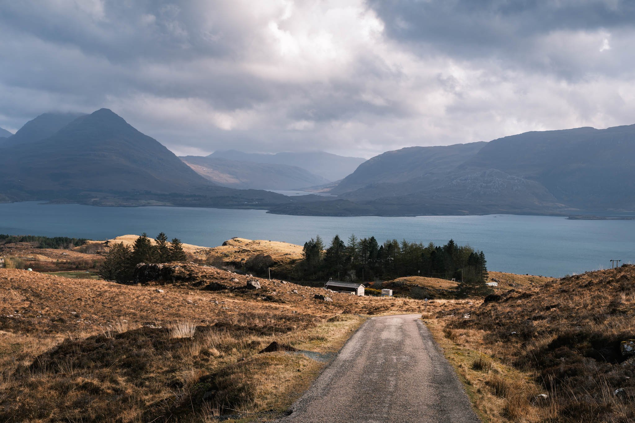 Highland - Scotland - Un chemin de terre mène à un Loch entouré de montagnes sous un ciel nuageux.