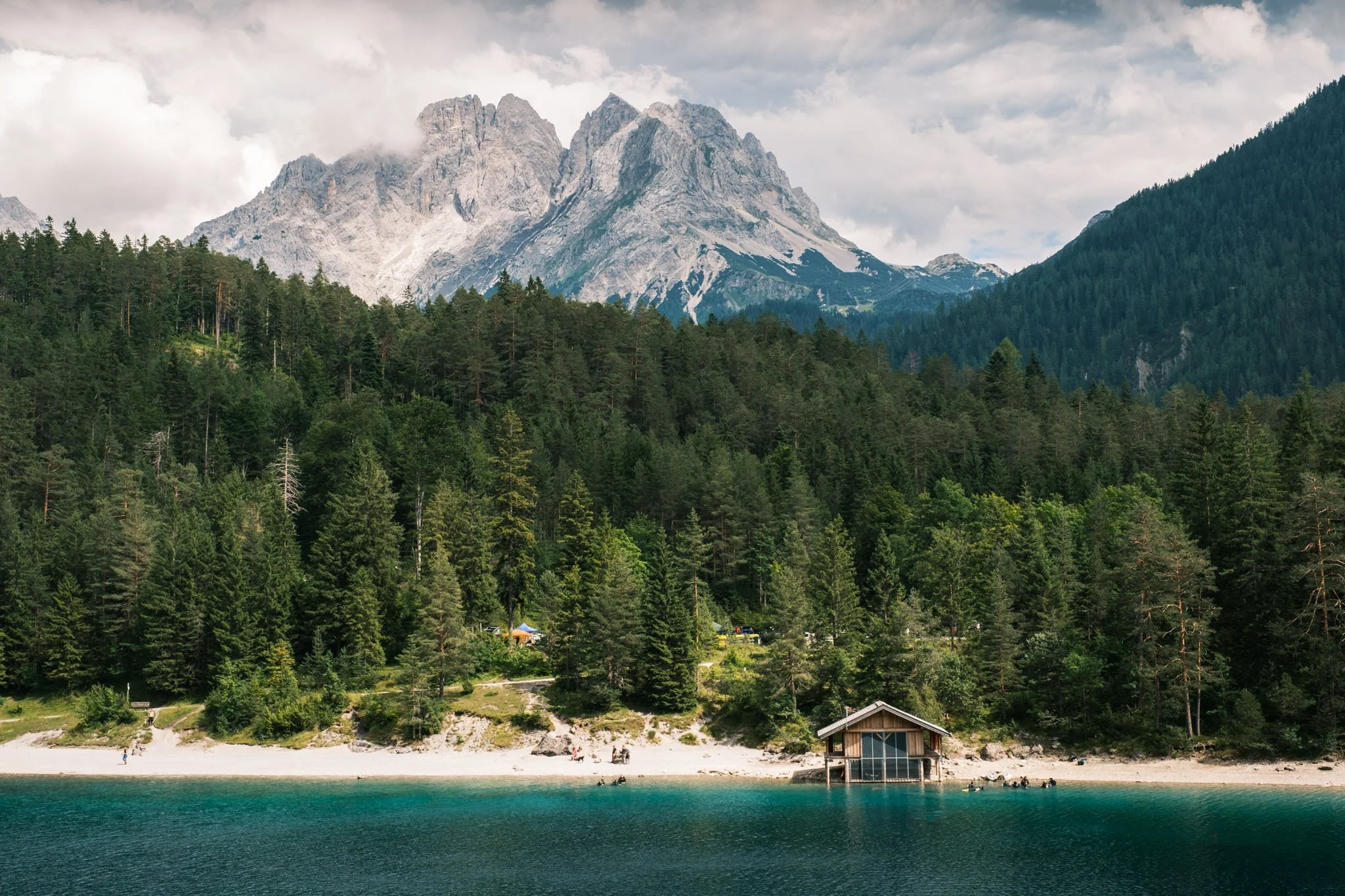 Tyrol - Lac avec une cabane en bois au bord de l'eau, forêt dense en arrière-plan, montagnes enneigées sous un ciel nuageux. Reportage outdoor, mise en valeur du territoire et tourisme durable par Guillaume Donsimoni