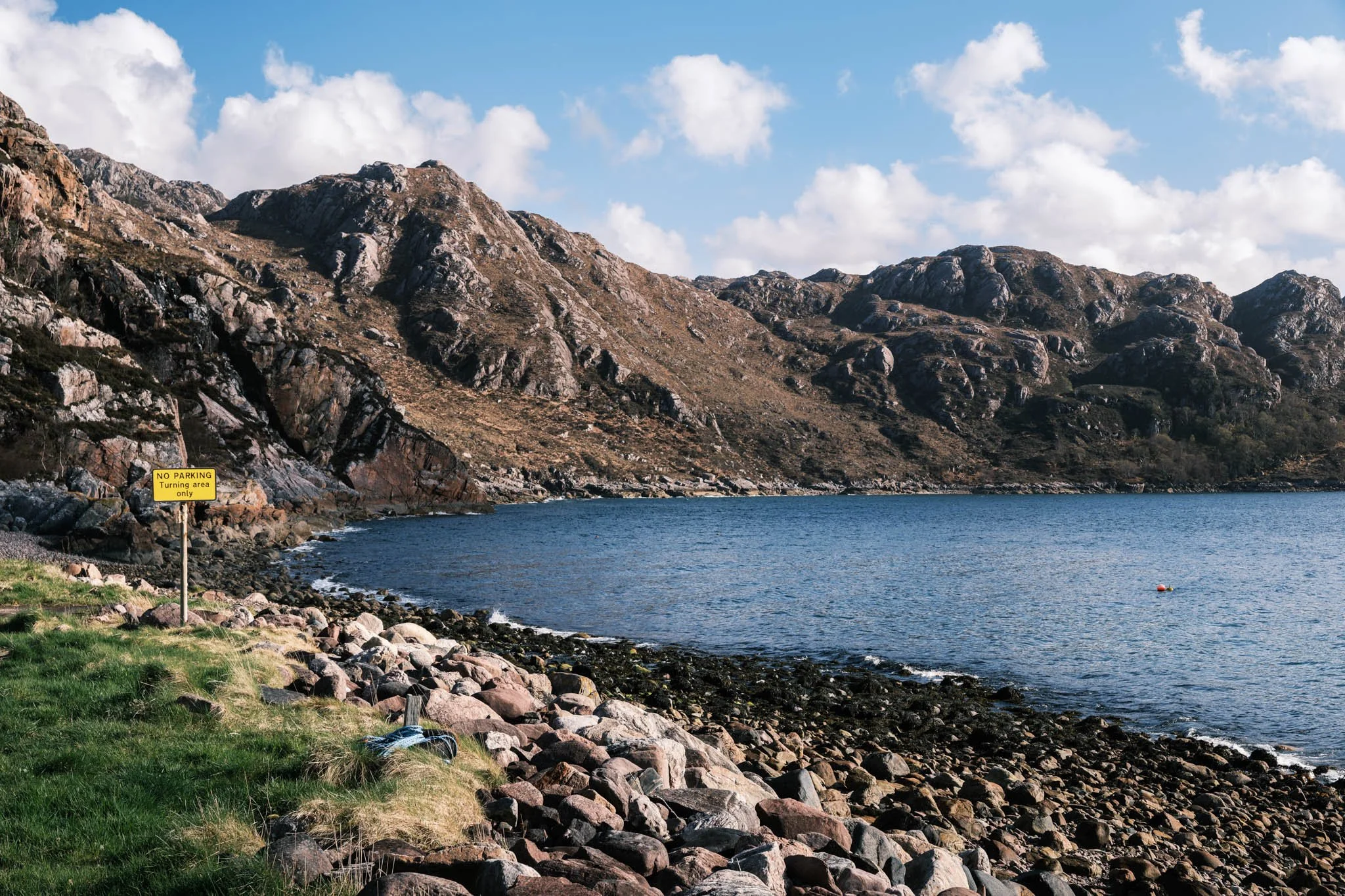 Highland - Scotland - Paysage de montagnes rocheuses avec un Loch au premier plan et un ciel bleu avec des nuages. Un panneau jaune indique 'NO PARKING Turning area only'.