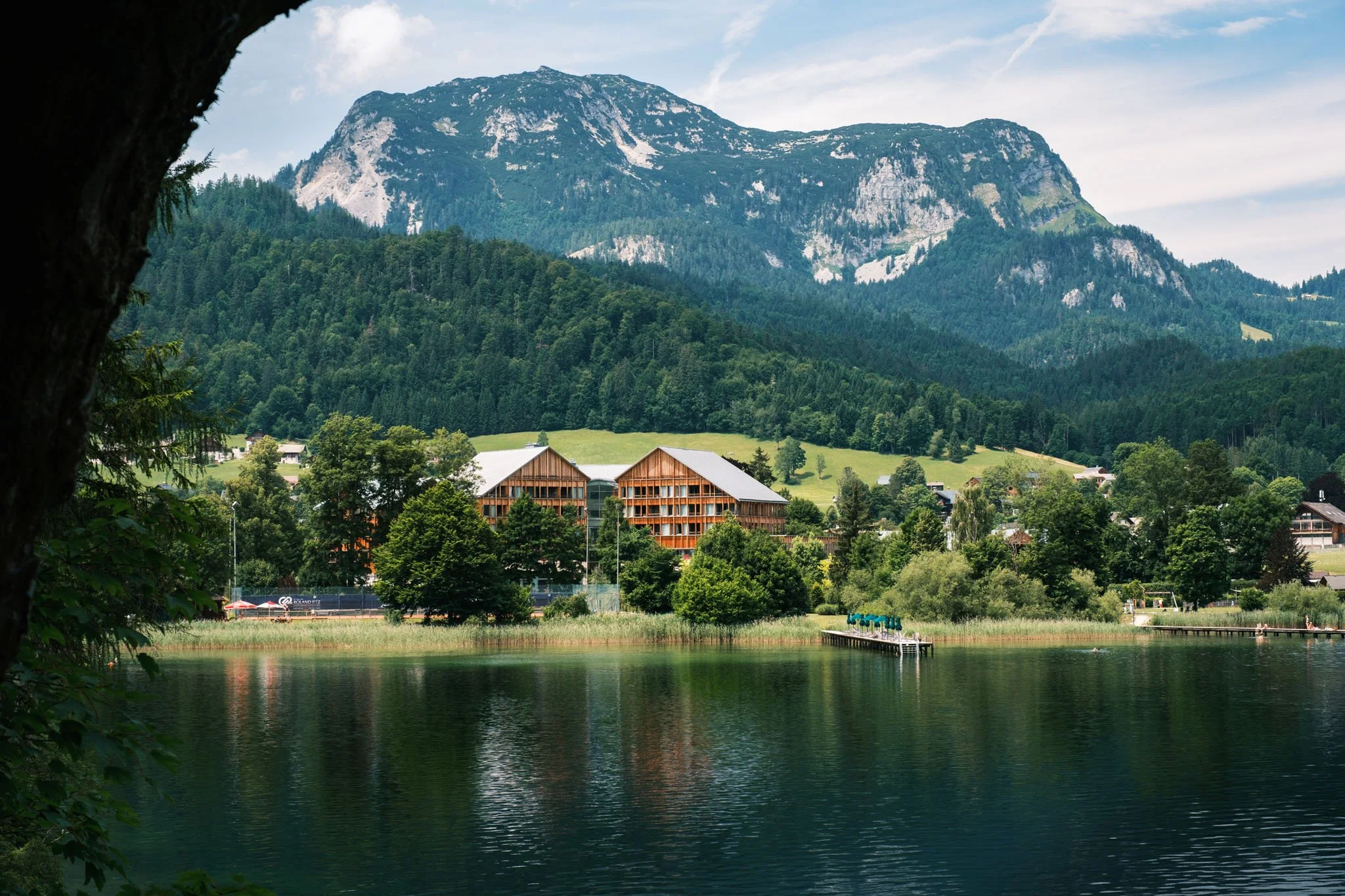 Austria - Vue d'un lac entouré d'arbres, avec des bâtiments en bois et une montagne en fond, sous un ciel partiellement nuageux. Reportage outdoor, mise en valeur du territoire et tourisme durable par Guillaume Donsimoni