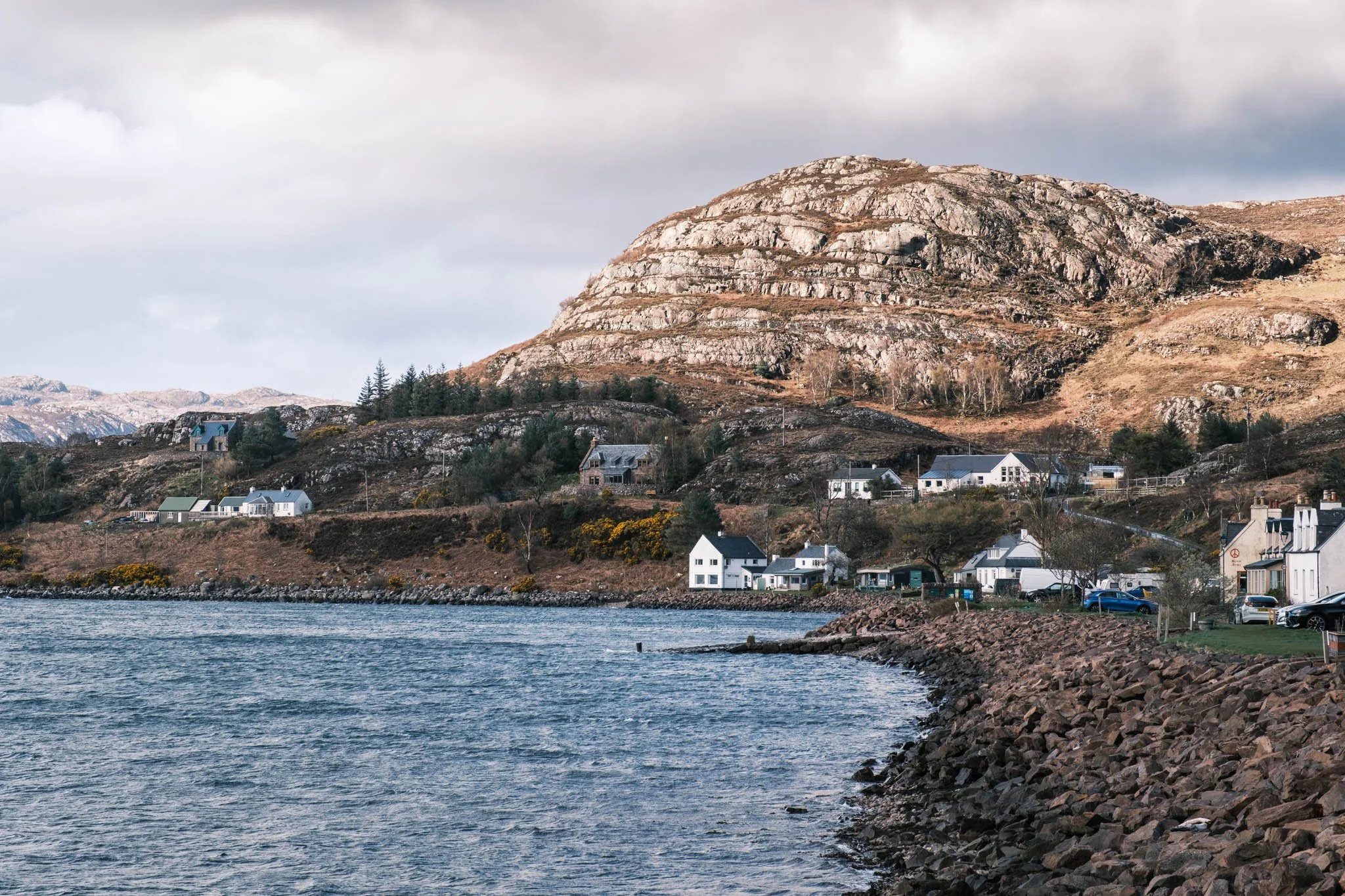 Highland - Scotland - Une ville au bord d'un Loch, avec des maisons blanches et des montagnes rocheuses en arrière-plan, sous un ciel nuageux.