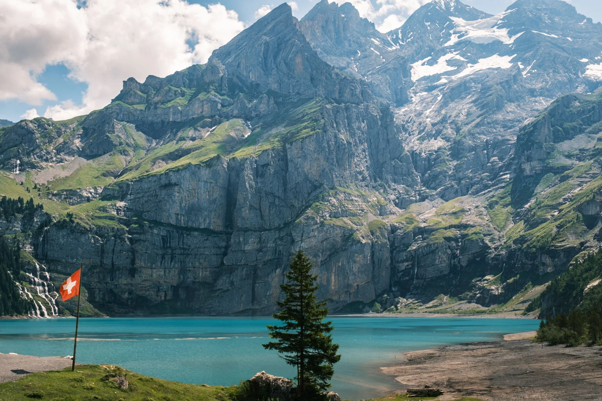 Oeschinensee. Lac turquoise entouré de montagnes rocheuses avec des sommets enneignés, un arbre et un drapeau suisse en premier plan. Reportage outdoor, mise en valeur du territoire et tourisme durable par Guillaume Donsimoni