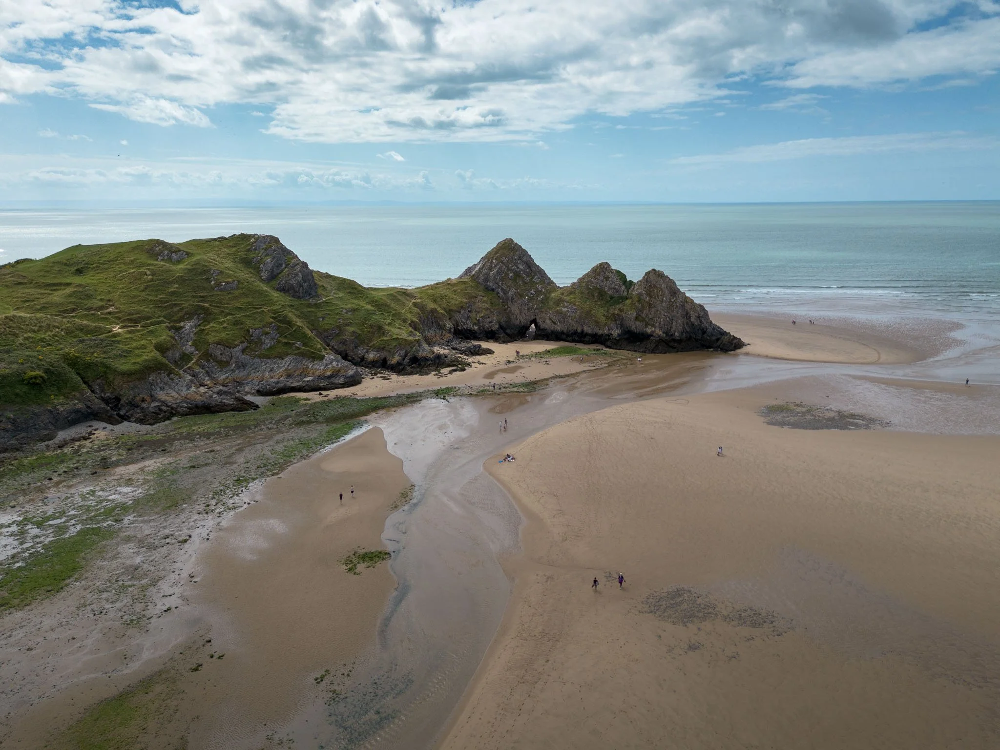 Sandy beach with green rocks in the background, sea view, partially cloudy sky, with a few people walking or sitting on the sand. Outdoor reportage, regional promotion, and sustainable tourism by Guillaume Donsimoni.
