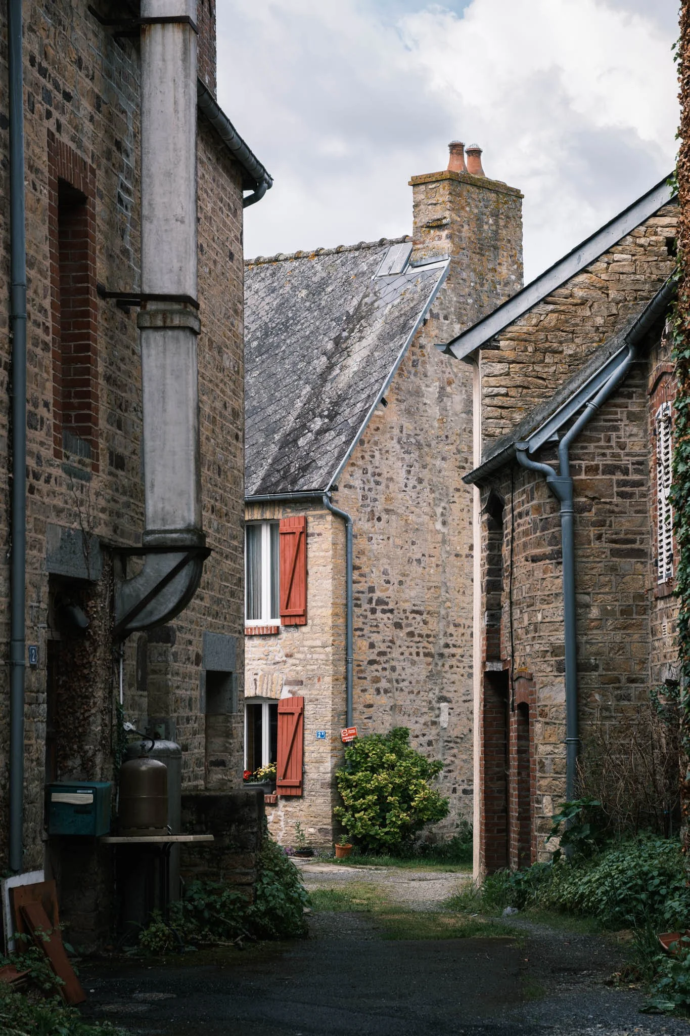 Suisse Normande - Une cour intérieure entourée de bâtiments en pierre avec des cheminées et des fenêtres à volets rouges, des plantes vertes autour, un ciel nuageux en arrière-plan.