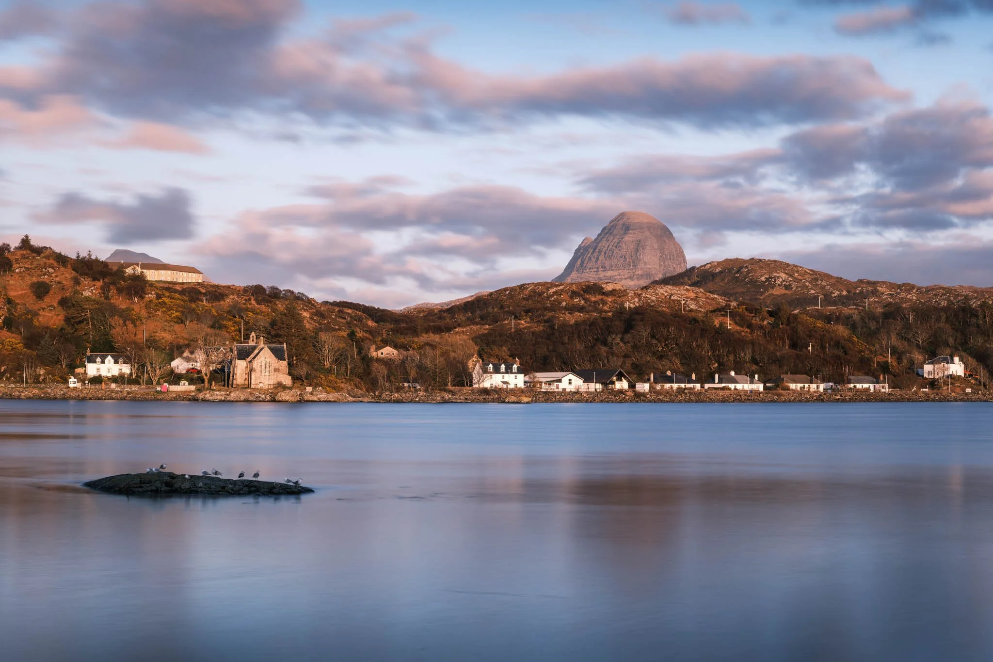 Highland - Scotland - Paysage côtier avec des maisons blanches au bord de l'eau, une colline avec des arbres et un grand rocher en forme de pyramide au loin, sous un ciel partiellement nuageux