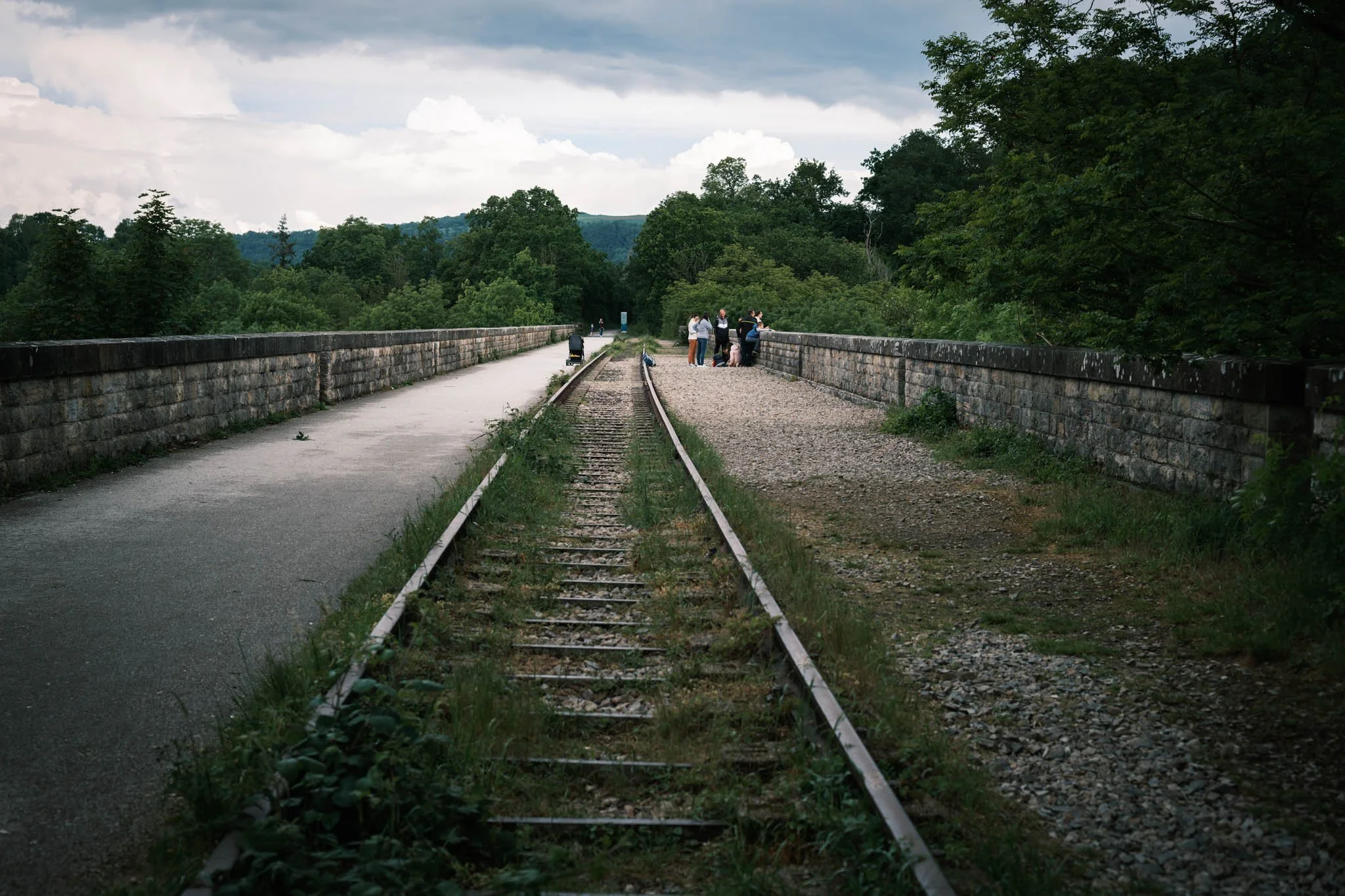 Suisse Normande - Vue d'un pont en pierre avec des rails de train abandonnés au milieu, entouré d'une verdure dense et de nuages gris dans le ciel.