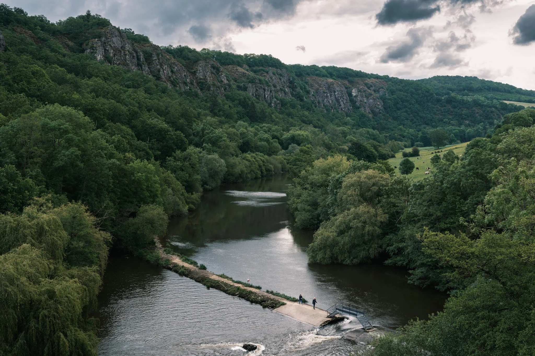 Suisse Normande - Deux personnes traversant une rivière sur un petit pont au milieu d'une forêt verte et d'une vallée entourée de collines rocheuses et de nuages dans le ciel.