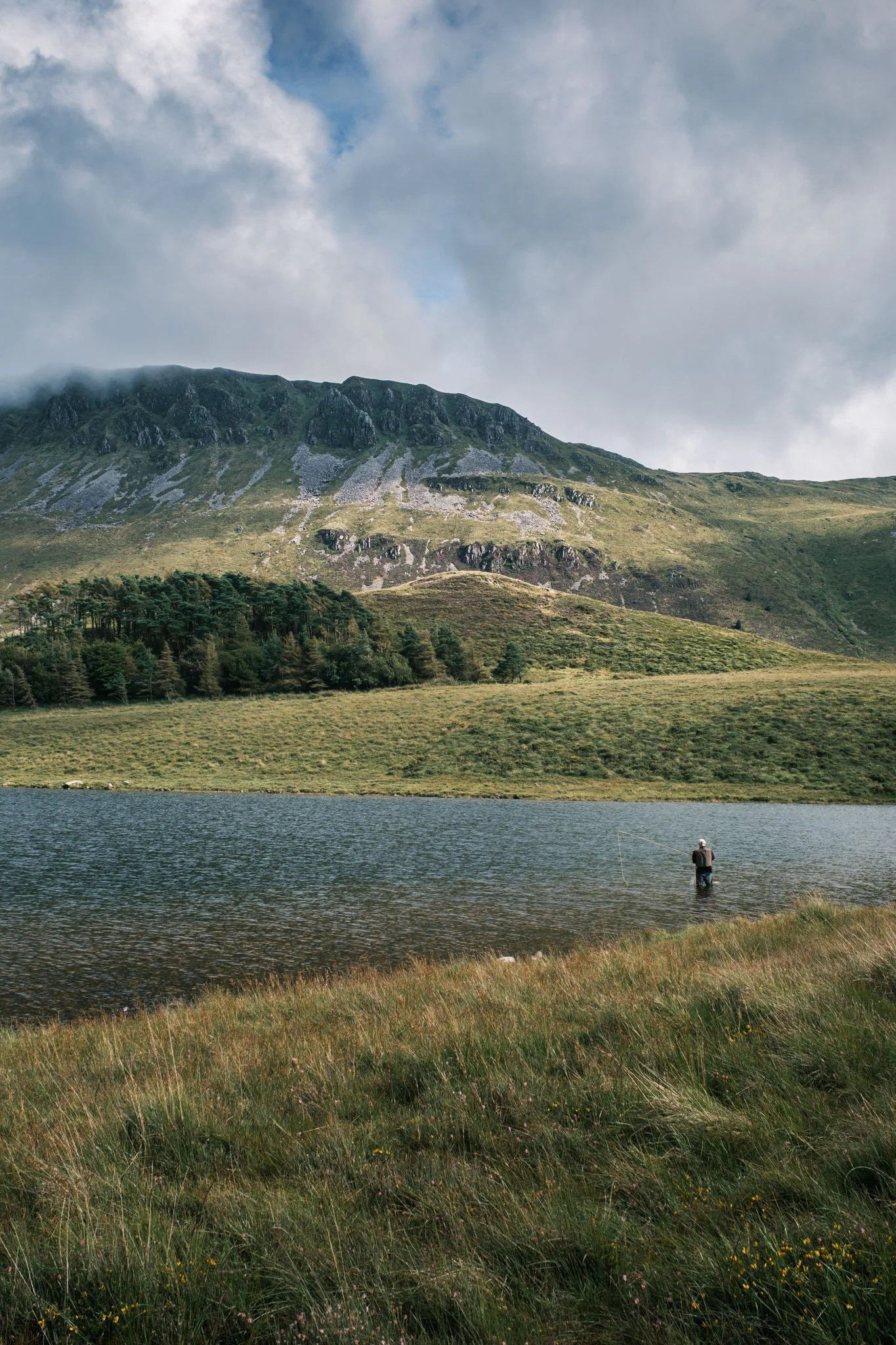 Wales - A fisherman standing in a lake surrounded by green hills and mountains under a cloudy sky.