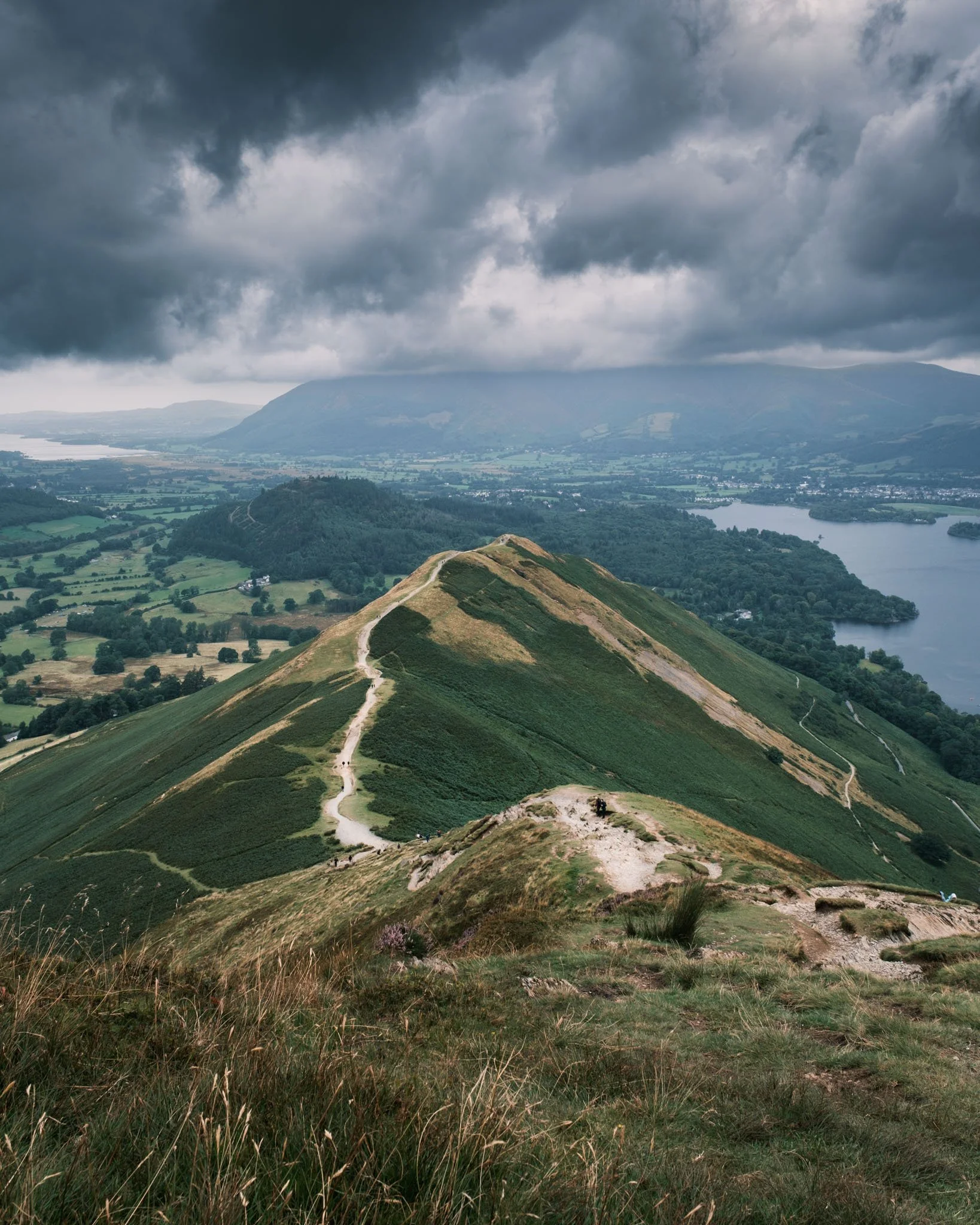 Lake District - Vue d'une montagne avec un sentier peu fréquenté, surplombant un paysage avec des lacs, des forêts et des champs, sous un ciel nuageux.  Reportage outdoor, mise en valeur du territoire et tourisme durable par Guillaume Donsimoni.