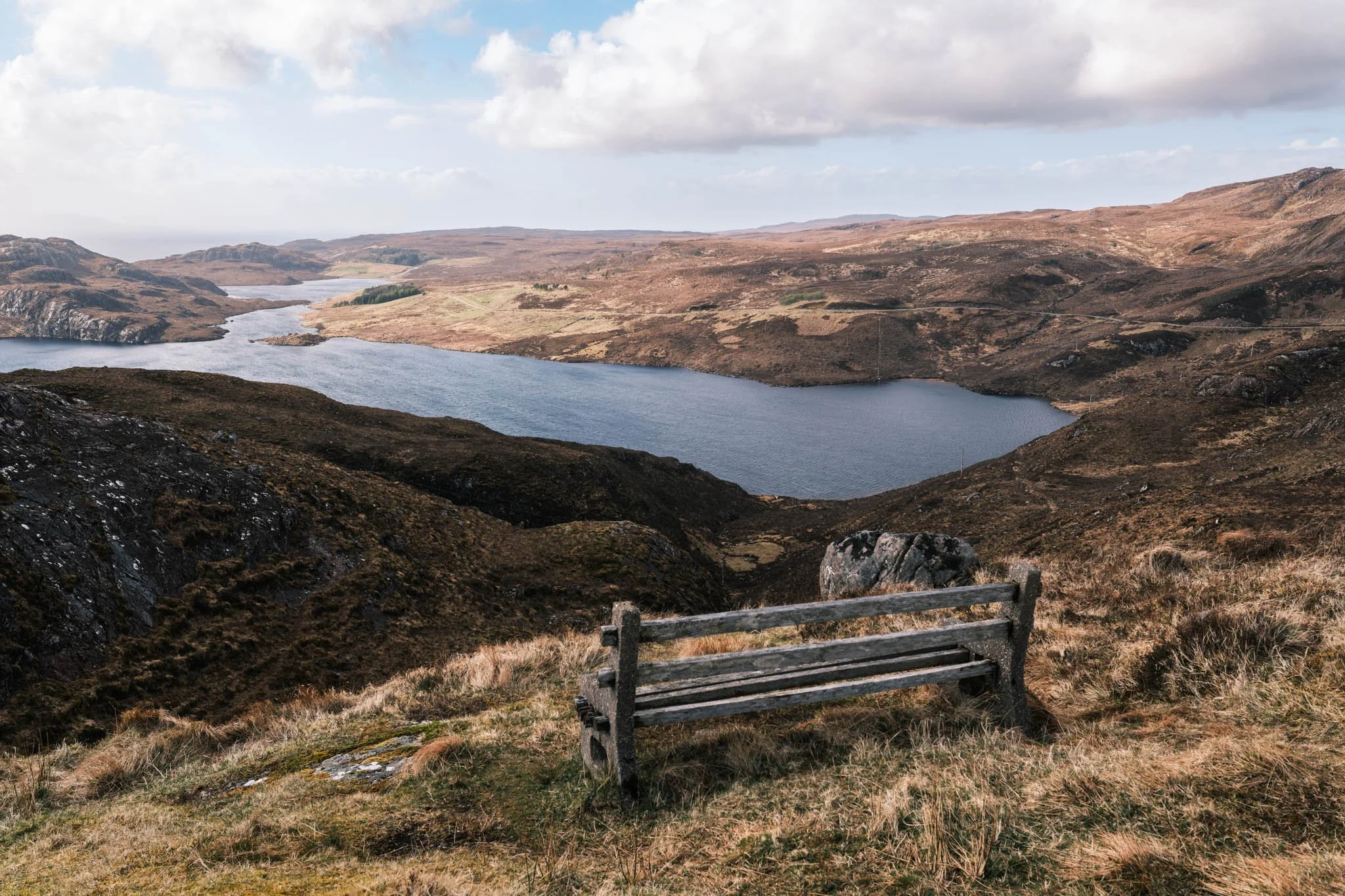 Highland - Scotland - Paysage de collines vallonnées avec un Loch, ciel partiellement nuageux, et un banc en bois sur un terrain herbeux.