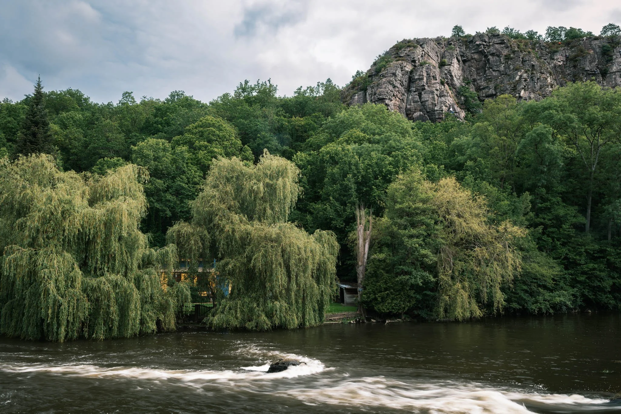 Suisse Normande - Une rivière entourée d'arbres verts avec une falaise rocheuse au fond, sous un ciel nuageux.