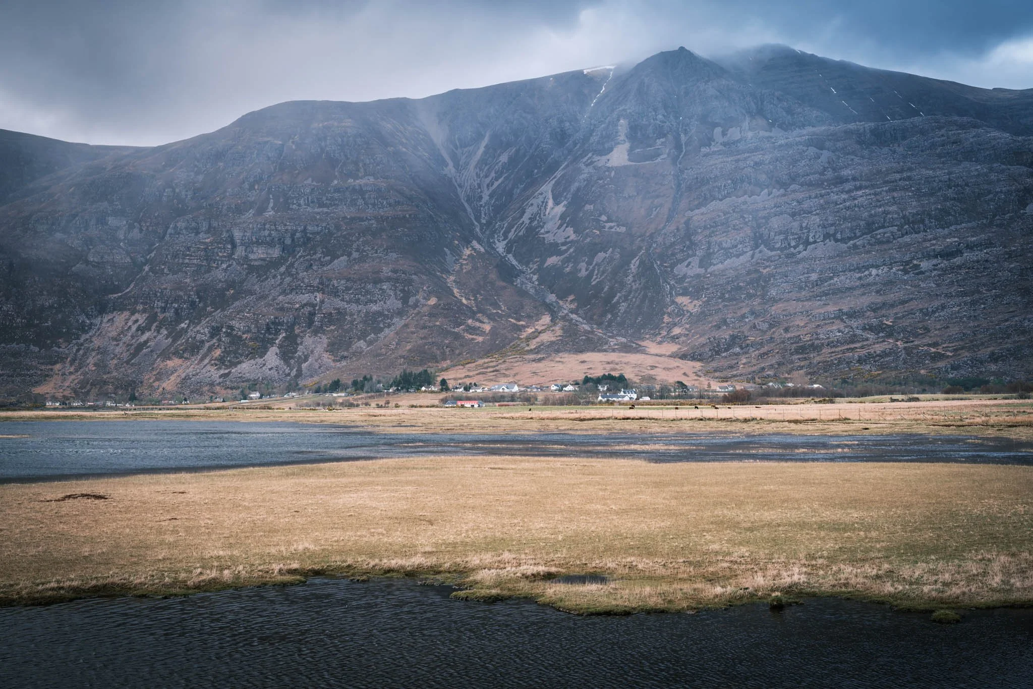 Highland - Scotland - Montagne imposante avec une petite ville à la base, rivière en premier plan et nuages au sommet.