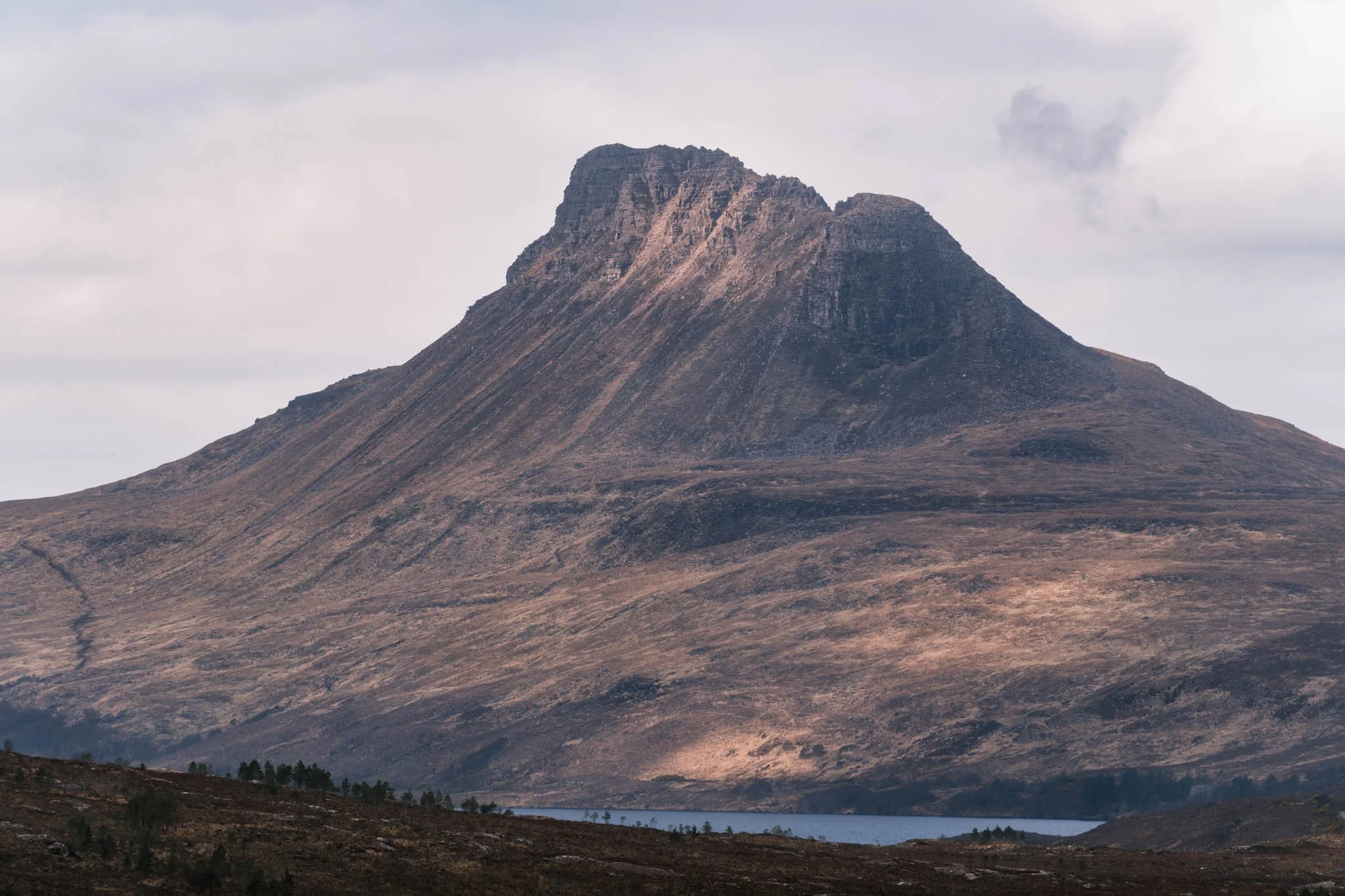 Highland - Scotland - Montagne rocheuse avec un sommet escarpé, terrain aride, Loch au premier plan, ciel nuageux.