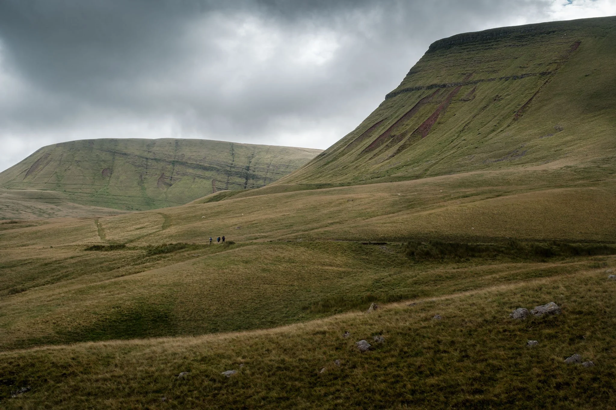 Wales: A landscape of verdant hills under a cloudy sky, with hikers traversing the terrain. Brecon Beacons. Outdoor photography, territorial promotion, and sustainable tourism by Guillaume Donsimoni.