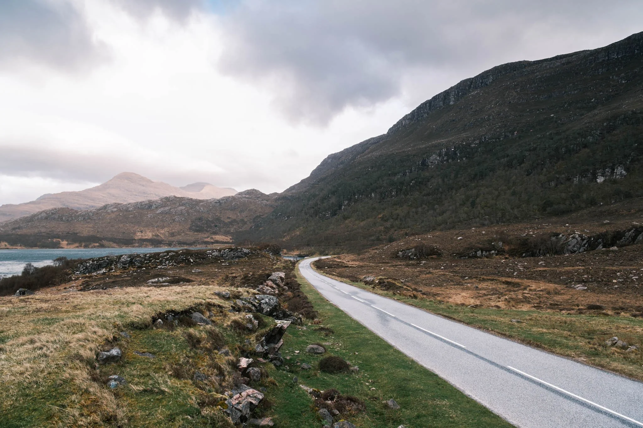 Highland - Scotland - Paysage de montagne avec une route sinueuse traversant une plaine verdoyante, sous un ciel nuageux.