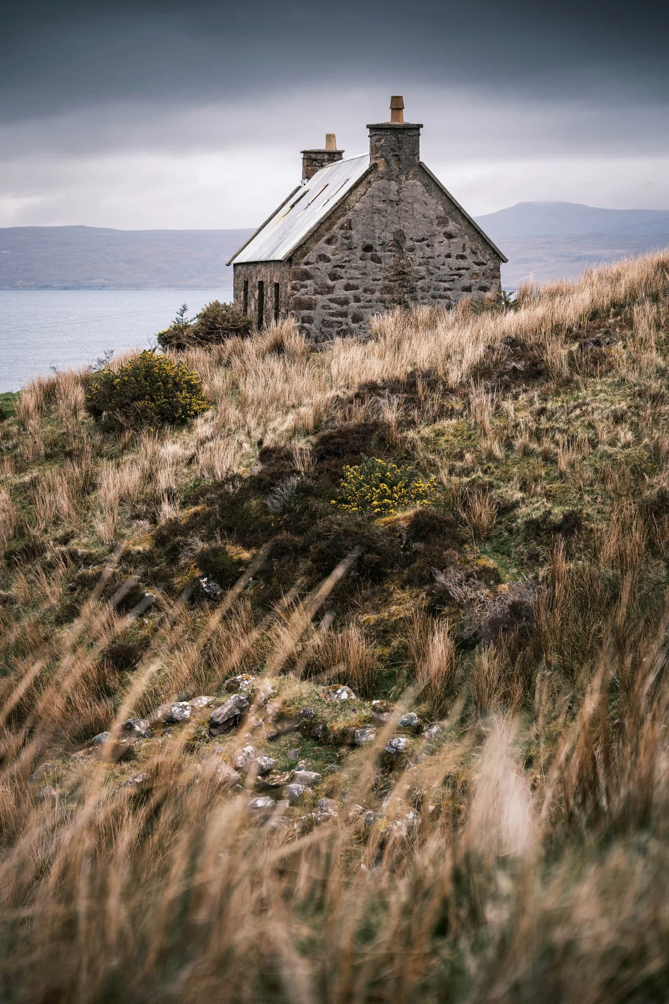 Highland - Scotland - Une maison en pierre au bord de l'eau avec un ciel nuageux, entourée d'herbes hautes et de buissons.