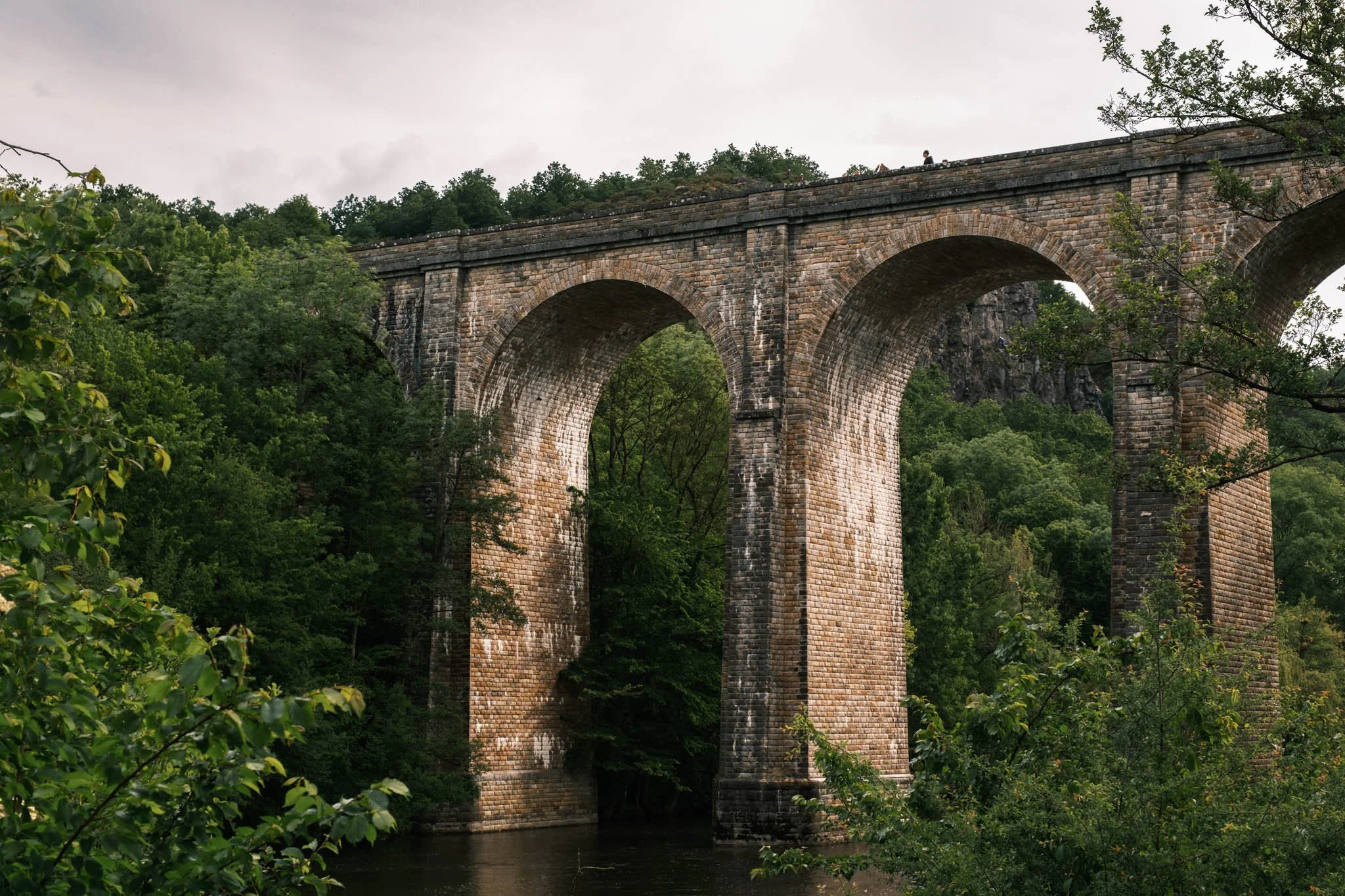 Suisse Normande - Un vieux pont en pierre traversant une rivière, entouré de verdure et de forêts, avec un ciel nuageux.