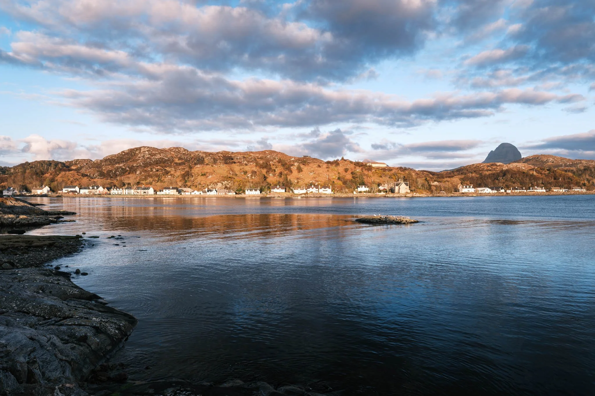Highland - Scotland - Une vue d'une côte avec des montagnes en arrière-plan, un ciel partiellement nuageux et de l'eau calme au premier plan, avec quelques maisons blanches au bord de la mer.