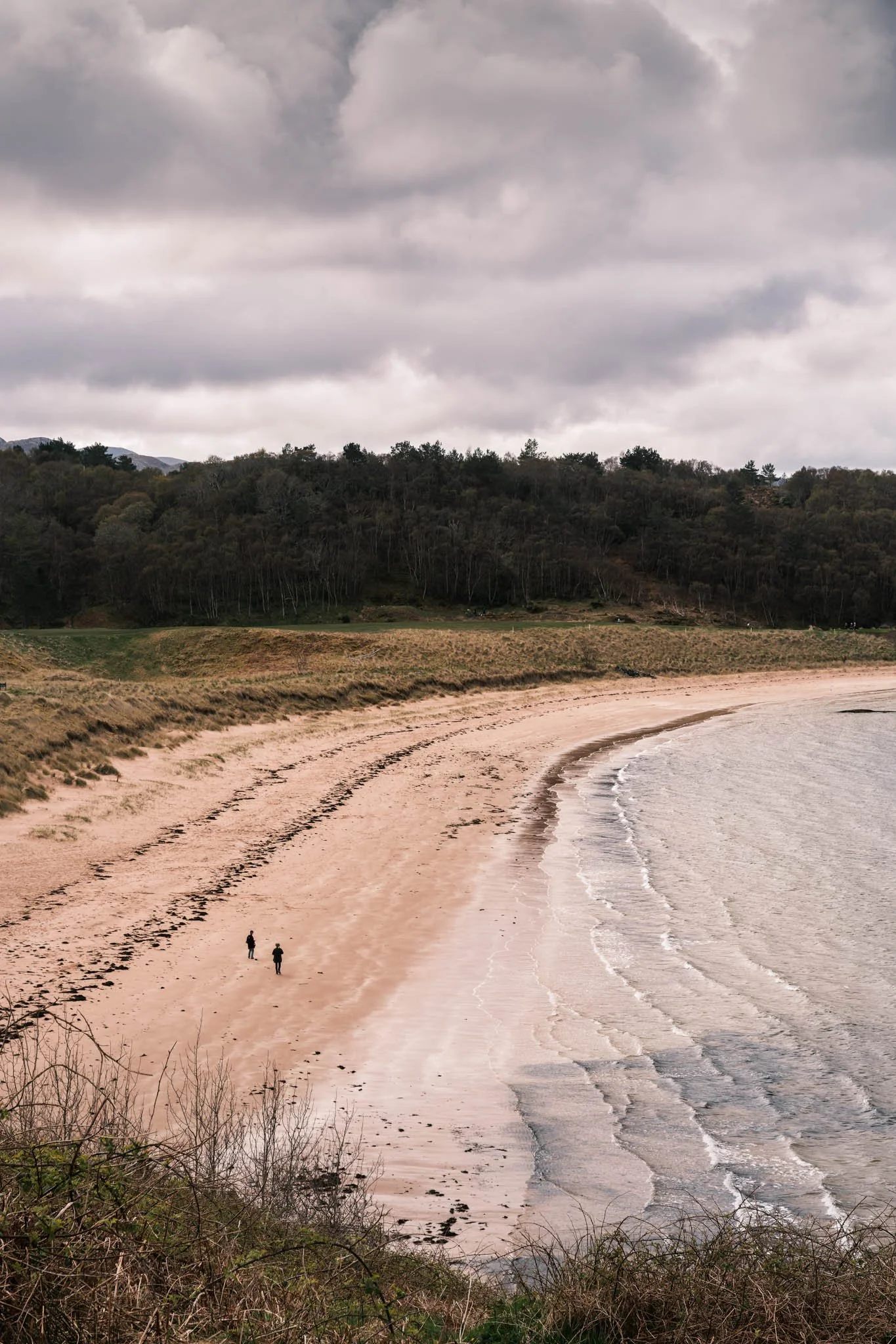 Highland - Scotland - Plage de sable avec deux personnes qui marchent, entourée de végétation et de collines verdoyantes, ciel nuageux.
