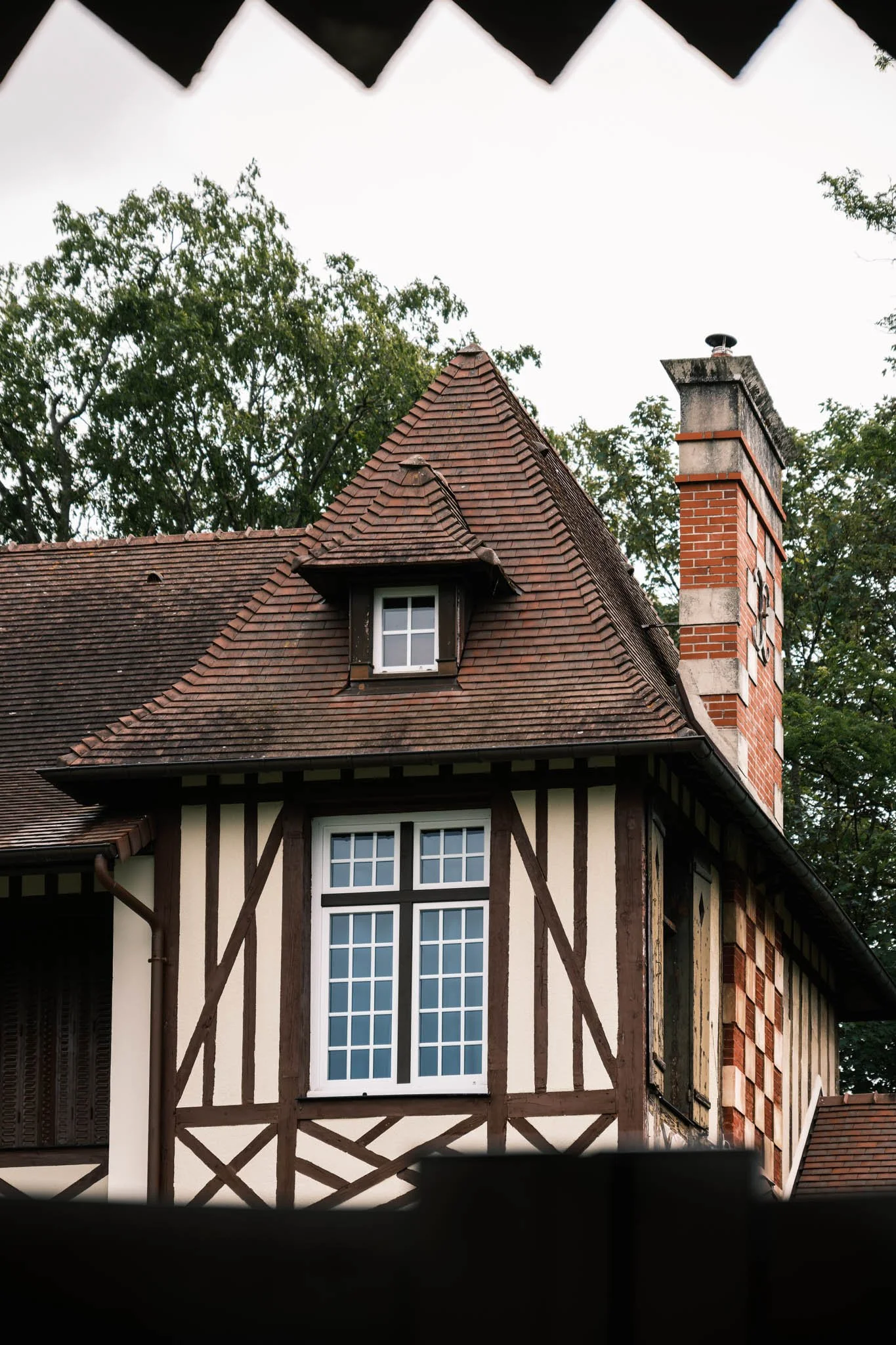 Suisse Normande - Maison à colombages avec toiture en tuiles rouges, cheminée en briques et fenêtres à petits carreaux, entourée d'arbres.