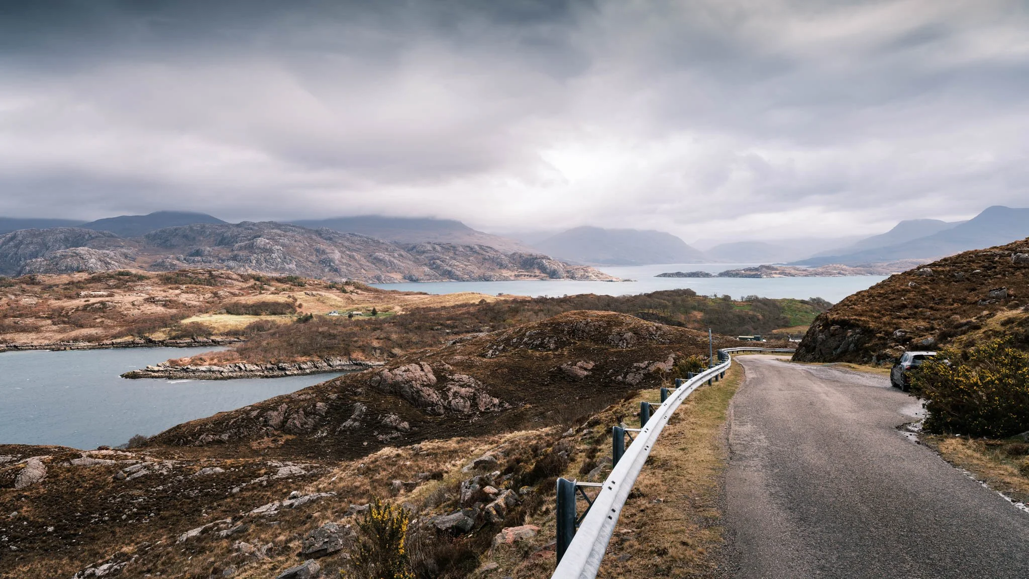 Highland - Scotland - Une route sinueuse bordée d'une barrière en métal, entourée de collines rocheuses et de végétation brune, avec un plan d'eau et des montagnes à l'horizon sous un ciel nuageux.