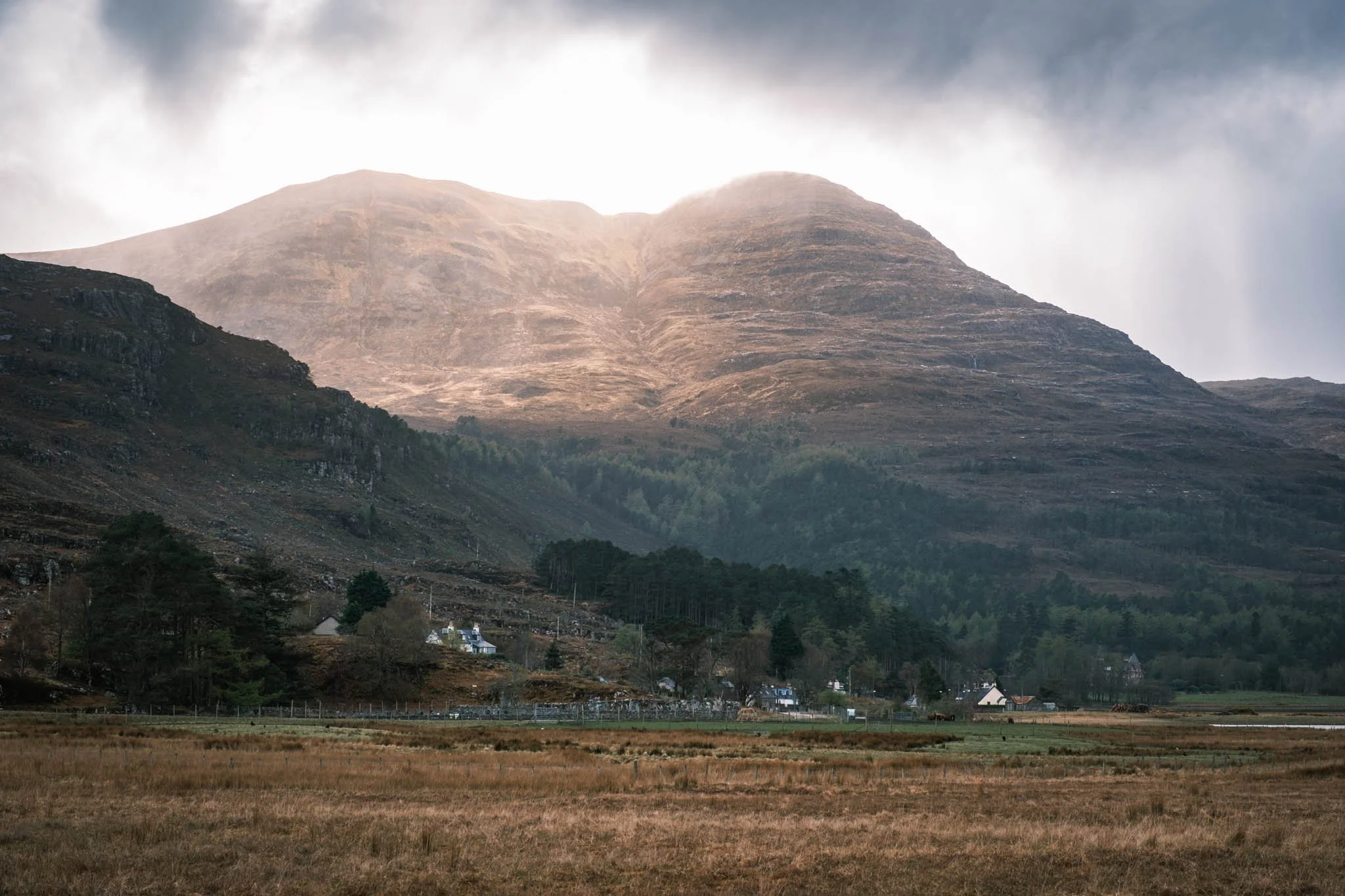 Highland - Scotland - Un paysage de montagne avec des collines verdoyantes et un petit village en bas, sous un ciel nuageux.