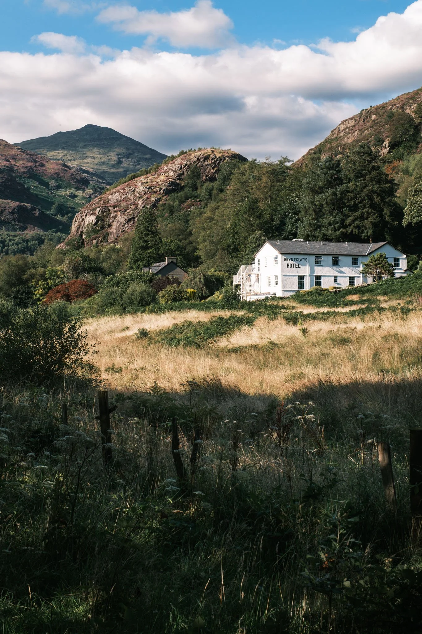 Wales - A white building situated in a mountainous landscape with green hills, trees, a partially cloudy sky, and a field of dry grass. Outdoor reportage, regional promotion, and sustainable tourism by Guillaum