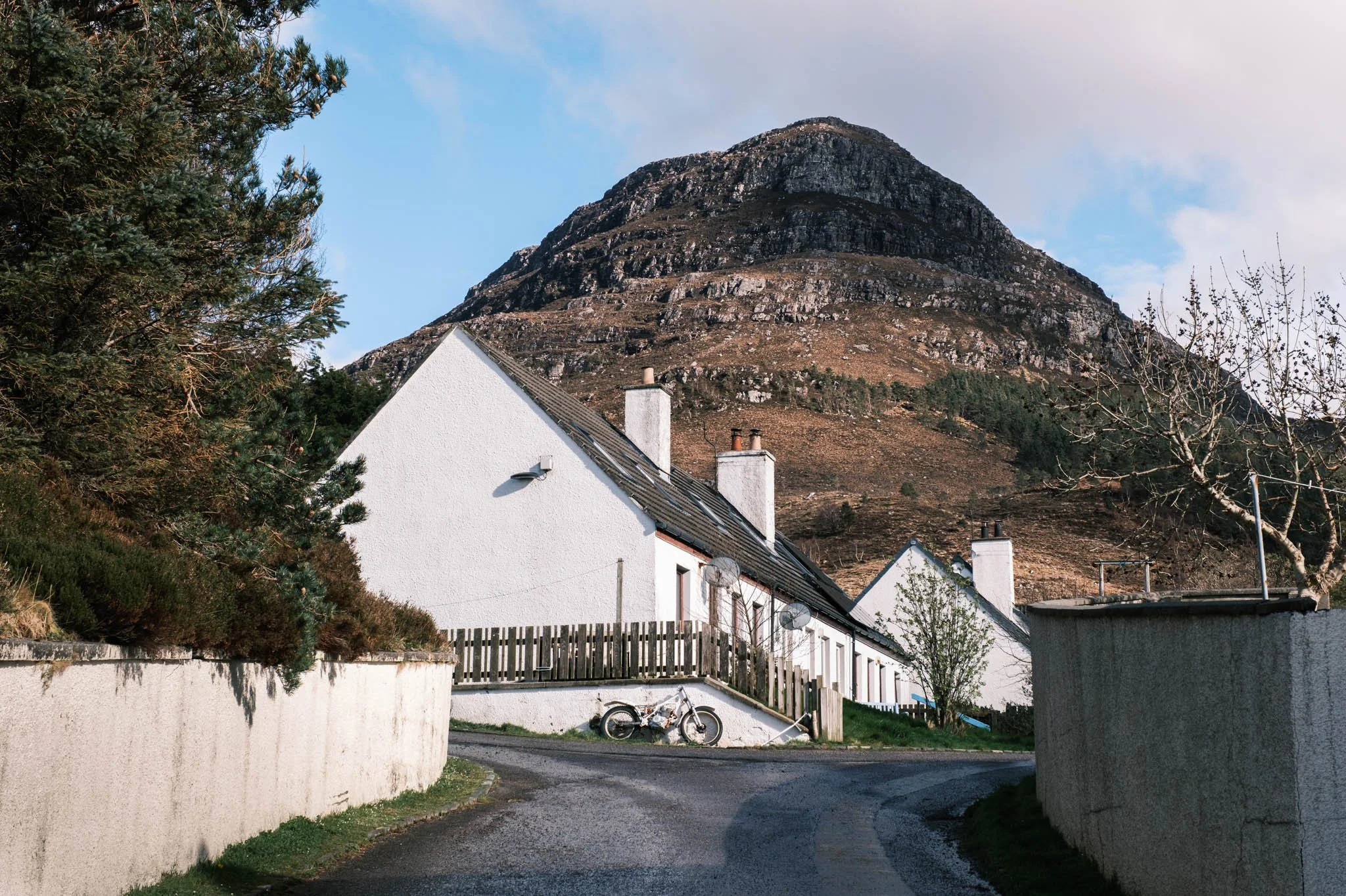 Highland - Scotland - Maisons blanches avec toits en tuiles sous une montagne rocheuse, avec quelques arbres et un vélo garé sur un chemin en premier plan.