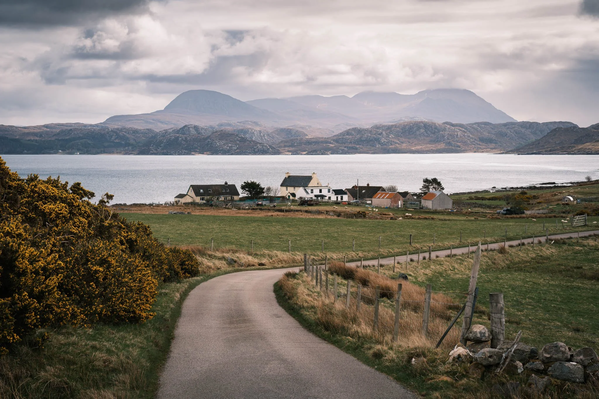 Highland - Scotland - Chemin de campagne serpentant vers des maisons rurales, avec un Loch et des montagnes en arrière-plan, sous un ciel nuageux.