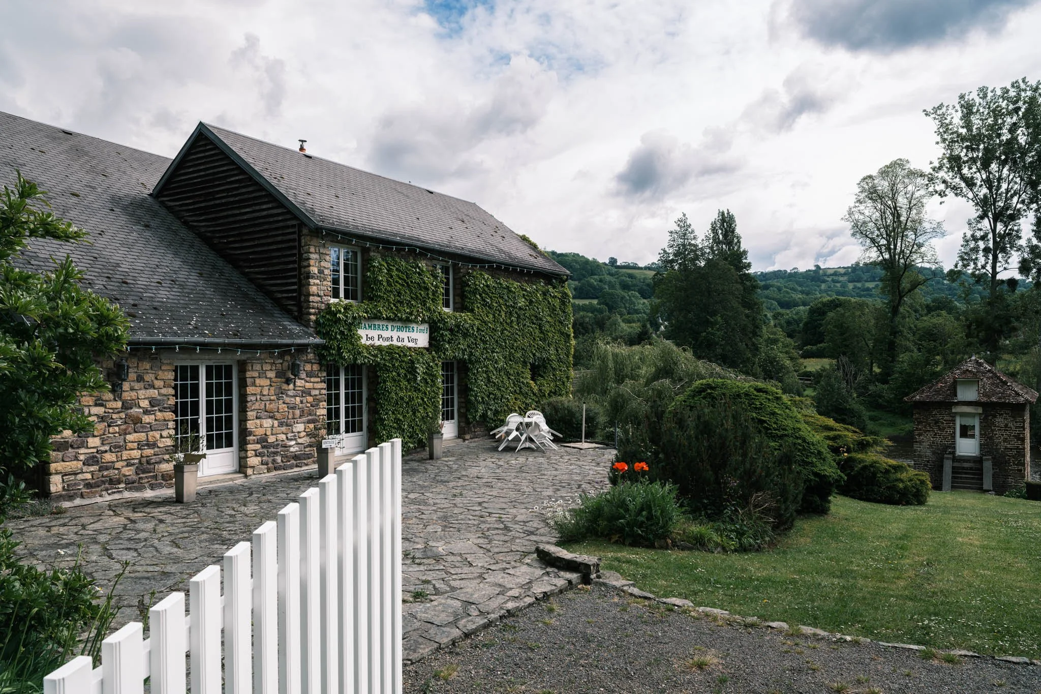 Suisse Normande - Une maison en pierre avec un toit en bardeaux sombre, entourée de beaucoup de verdure et de arbres, avec un ciel nuageux au-dessus.