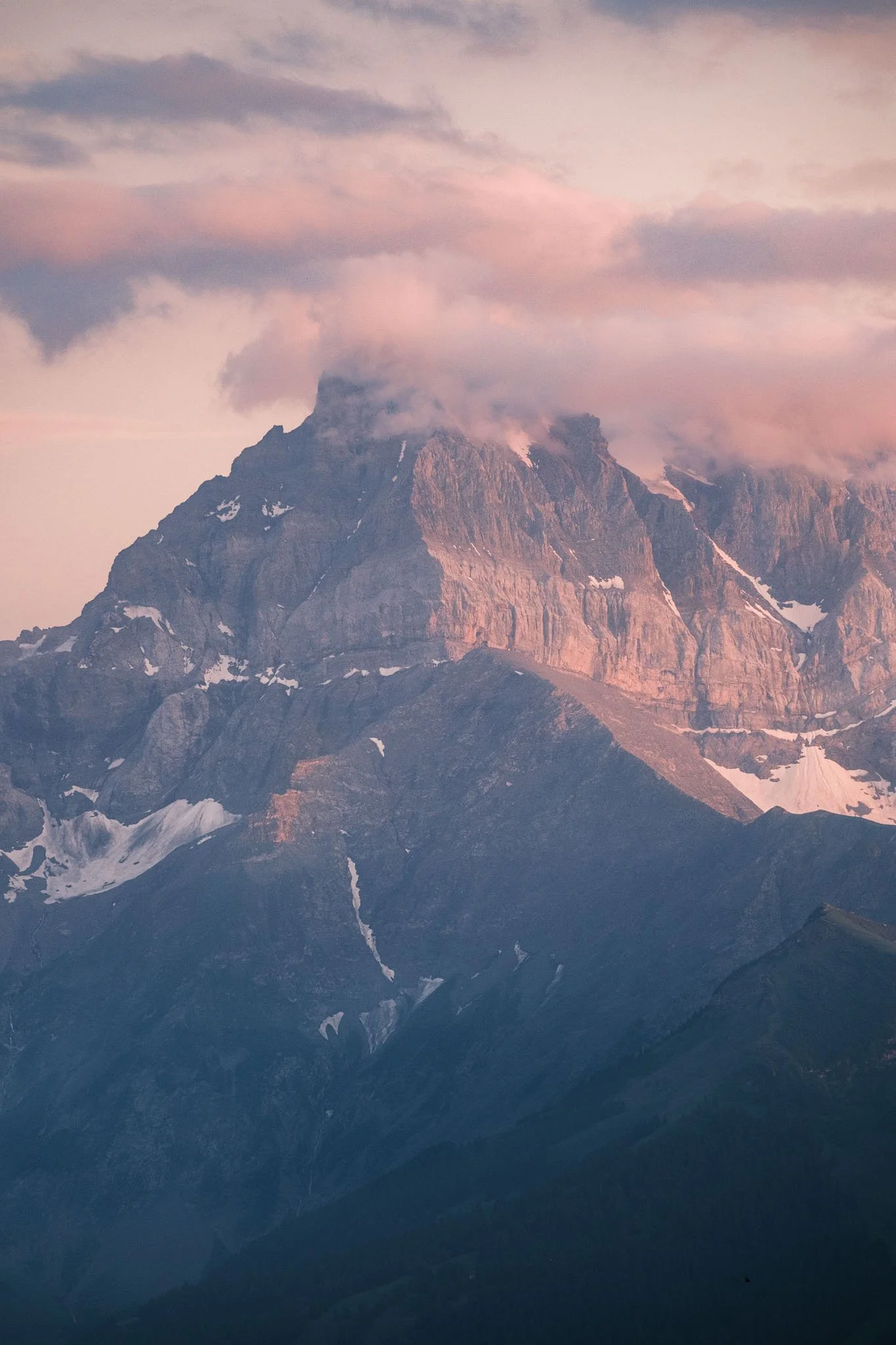 Une vue de montagnes rocheuses avec des rochers enneigés, partiellement couvertes de nuages, durant le coucher ou lever du soleil, avec un ciel pastel. Reportage outdoor, mise en valeur du territoire et tourisme durable par Guillaume Donsimoni