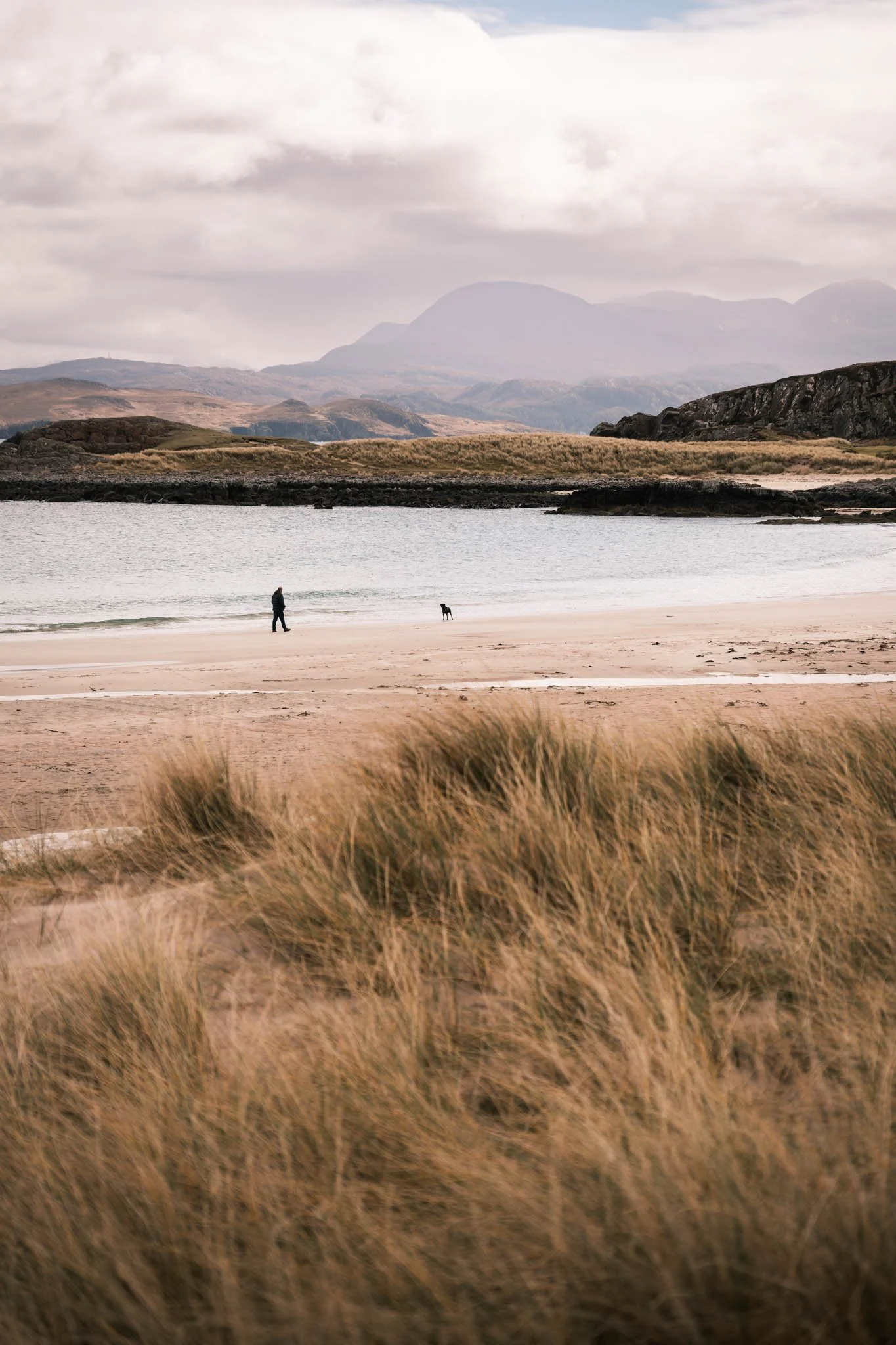 Highland - Scotland - Un homme et un chien marchant sur une plage entourée de dunes et de montagnes en arrière-plan, sous un ciel nuageux.