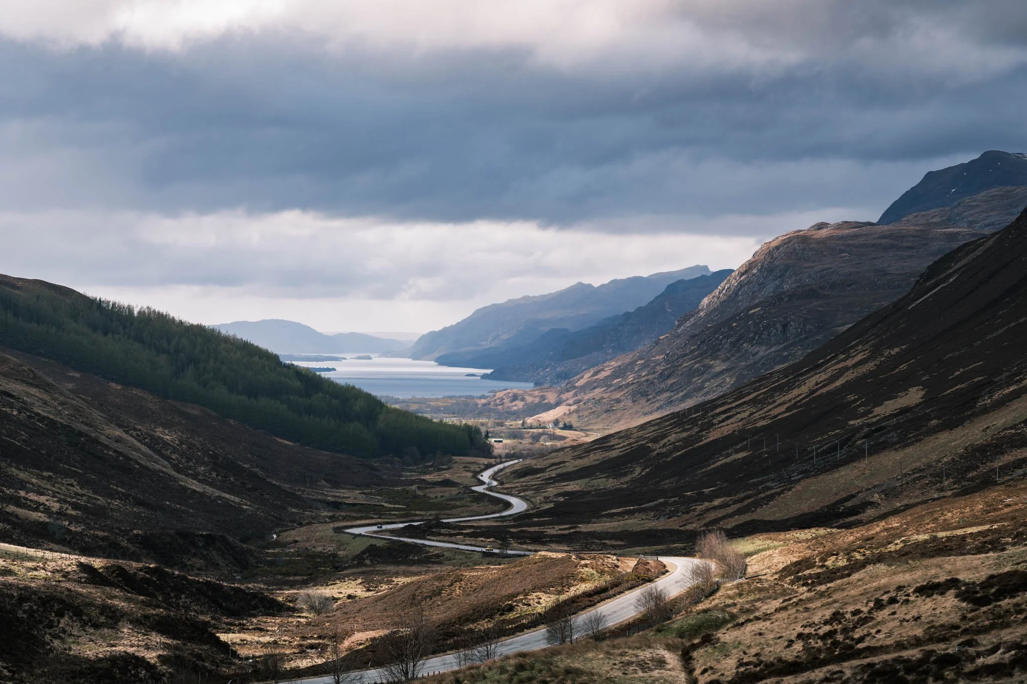 Highland - Scotland - Vallée montagneuse avec un Loch au fond, routes sinueuses, nuages gris et montagnes escarpées.